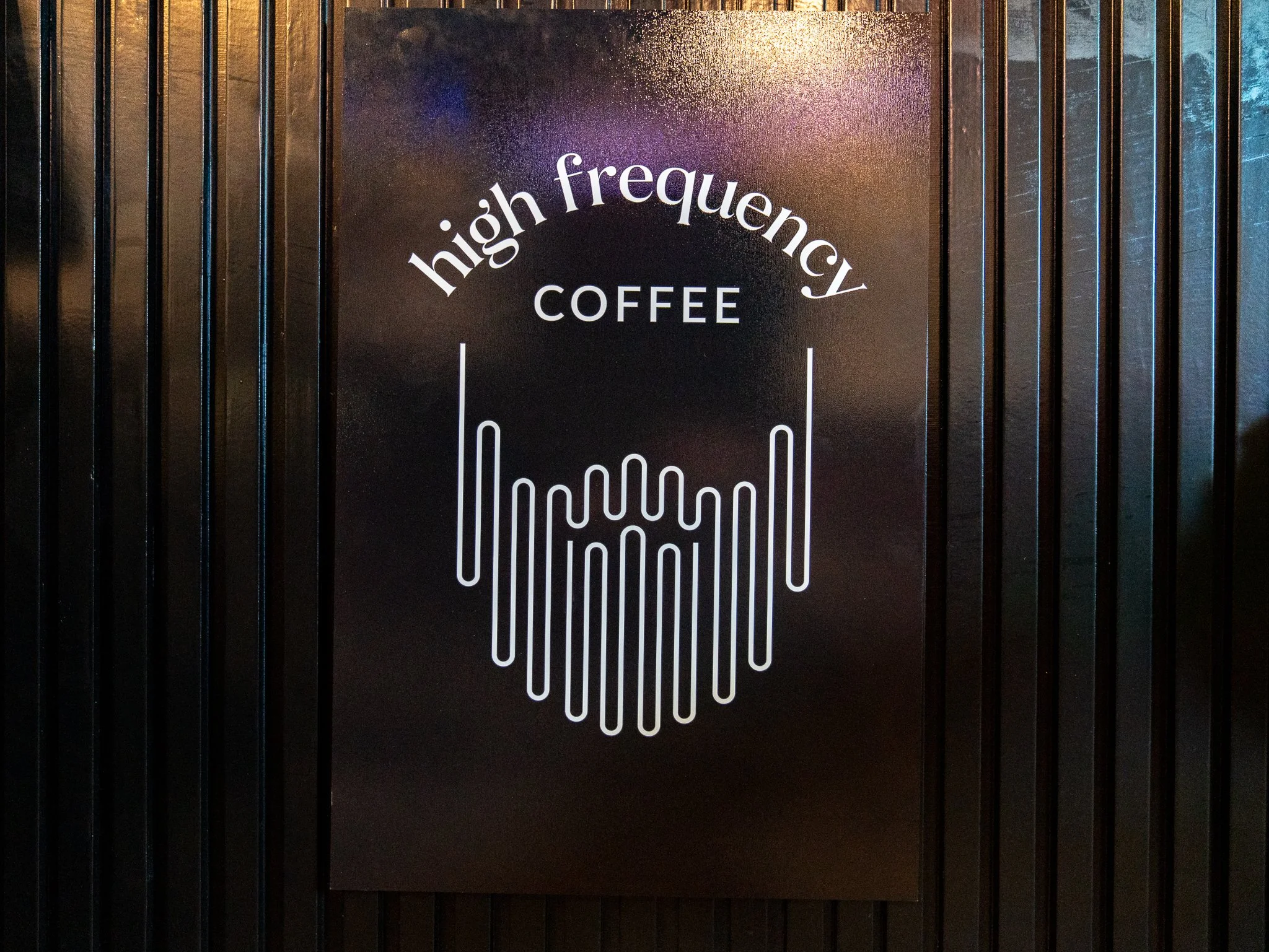 A black wall with a custom sign that reads 'high frequency COFFEE' in white text, featuring a stylized coffee cup formed with vertical lines.
