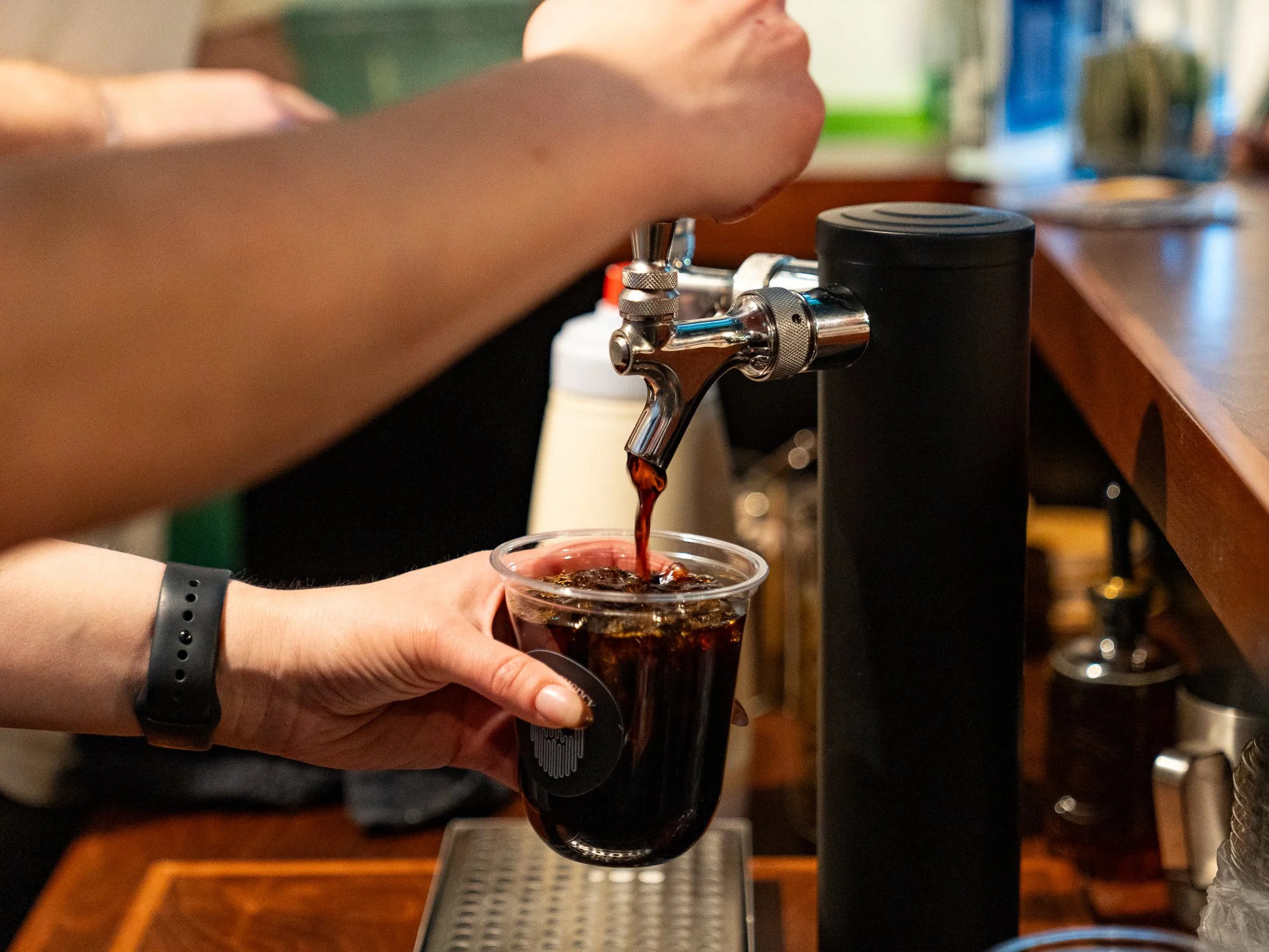 Barista pours cold brew coffee into a clear plastic cup from a tap at a bar or cafe. There a custom logo on the cup.