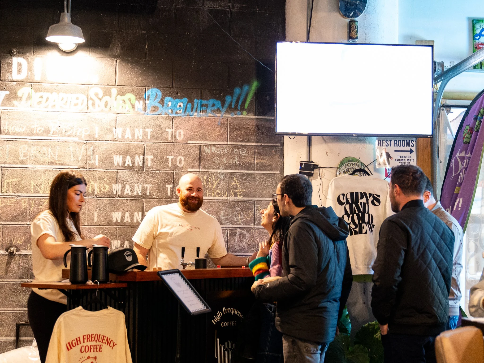 People standing in line at a coffee shop counter with a barista serving customers. A large TV screen is mounted on the wall behind them. The cafe has a chalkboard wall with writing and colorful graffiti in the background. One person behind the counte