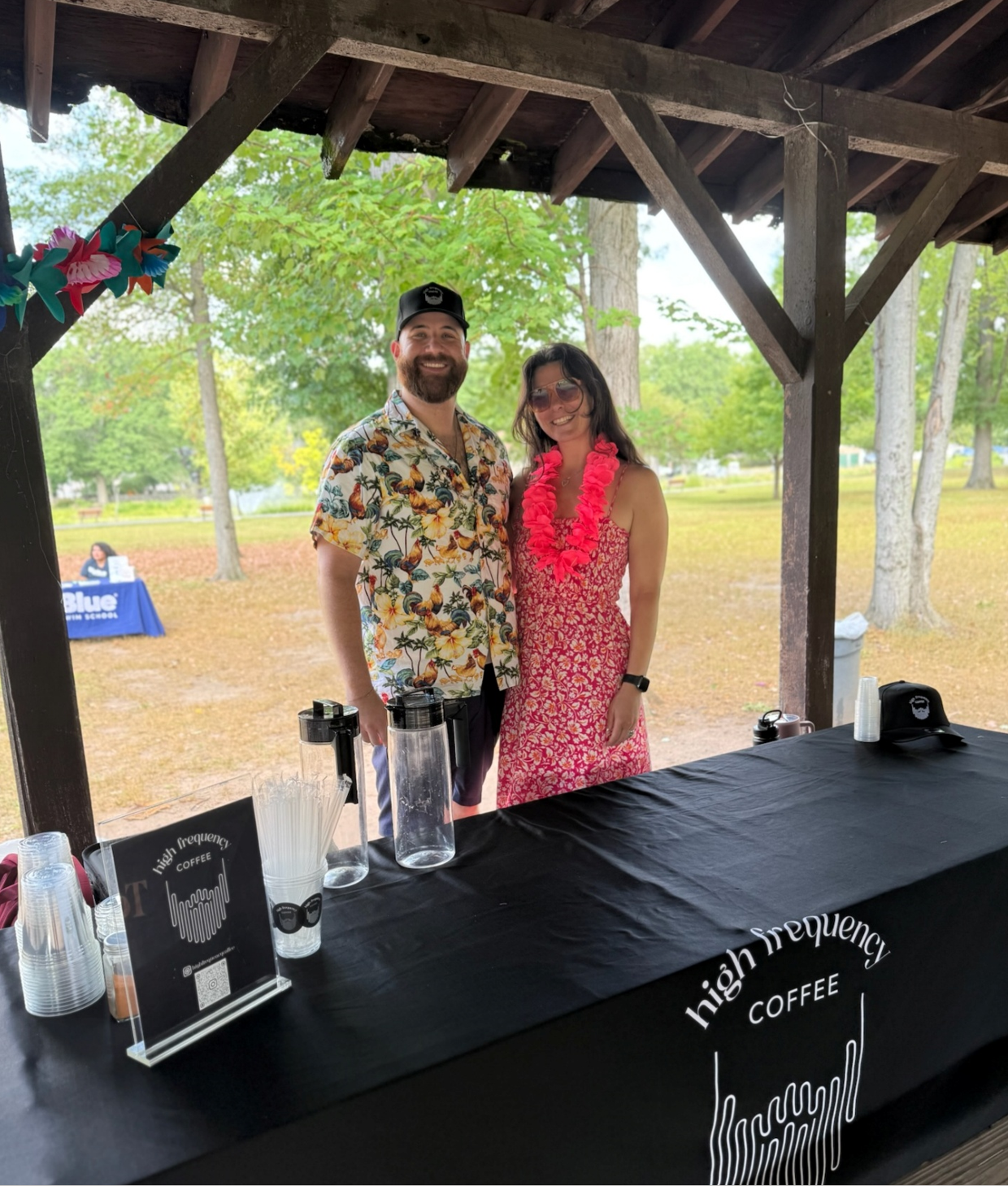 A smiling man and woman standing under a wooden pavilion at an outdoor event, with a table marked 'High Frequency Coffee' in front of them. The woman is wearing sunglasses and a red floral dress with a pink lei, the man is wearing a Hawaiian shirt and a cap. Trees and a park are visible in the background.