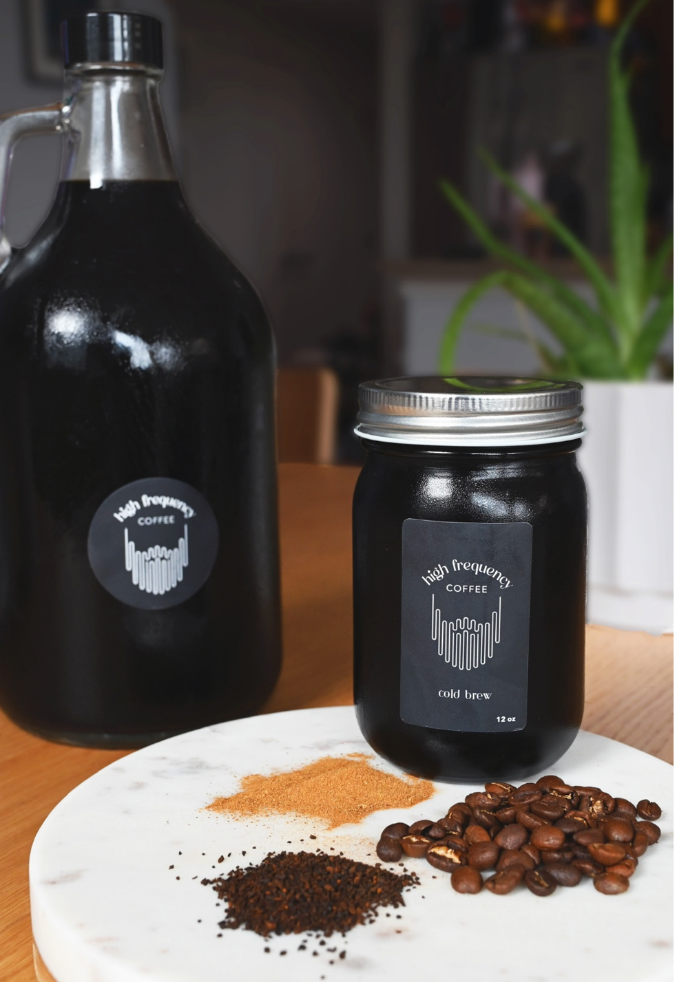 A jar and a bottle labeled "high frequency coffee cold brew" on a wooden table, with coffee beans and coffee grounds on a white marble surface, and a potted plant in the background.