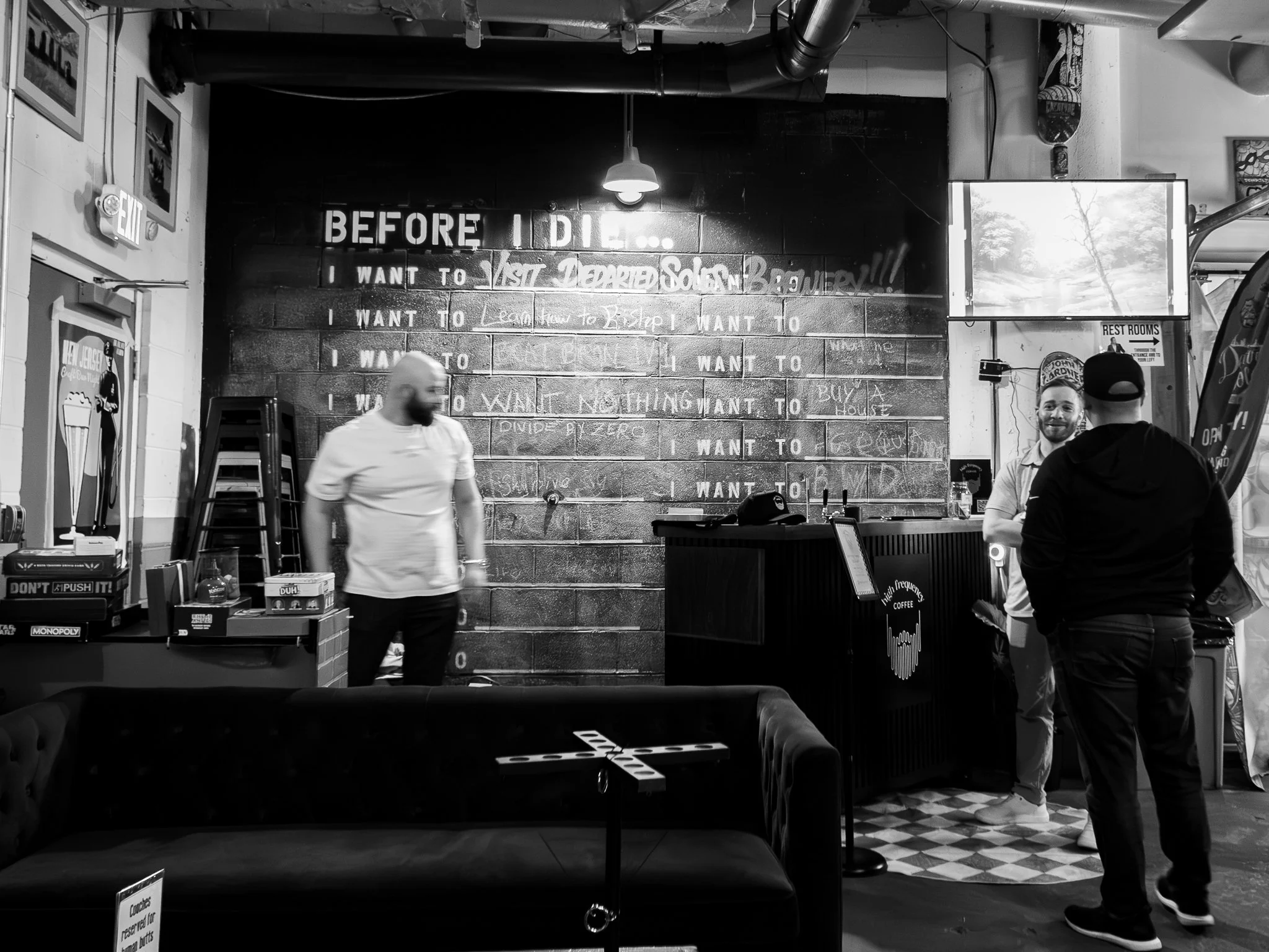 Inside a coffee shop with three men, one behind the counter and two customers. Chalkboard menu on the wall in the background.