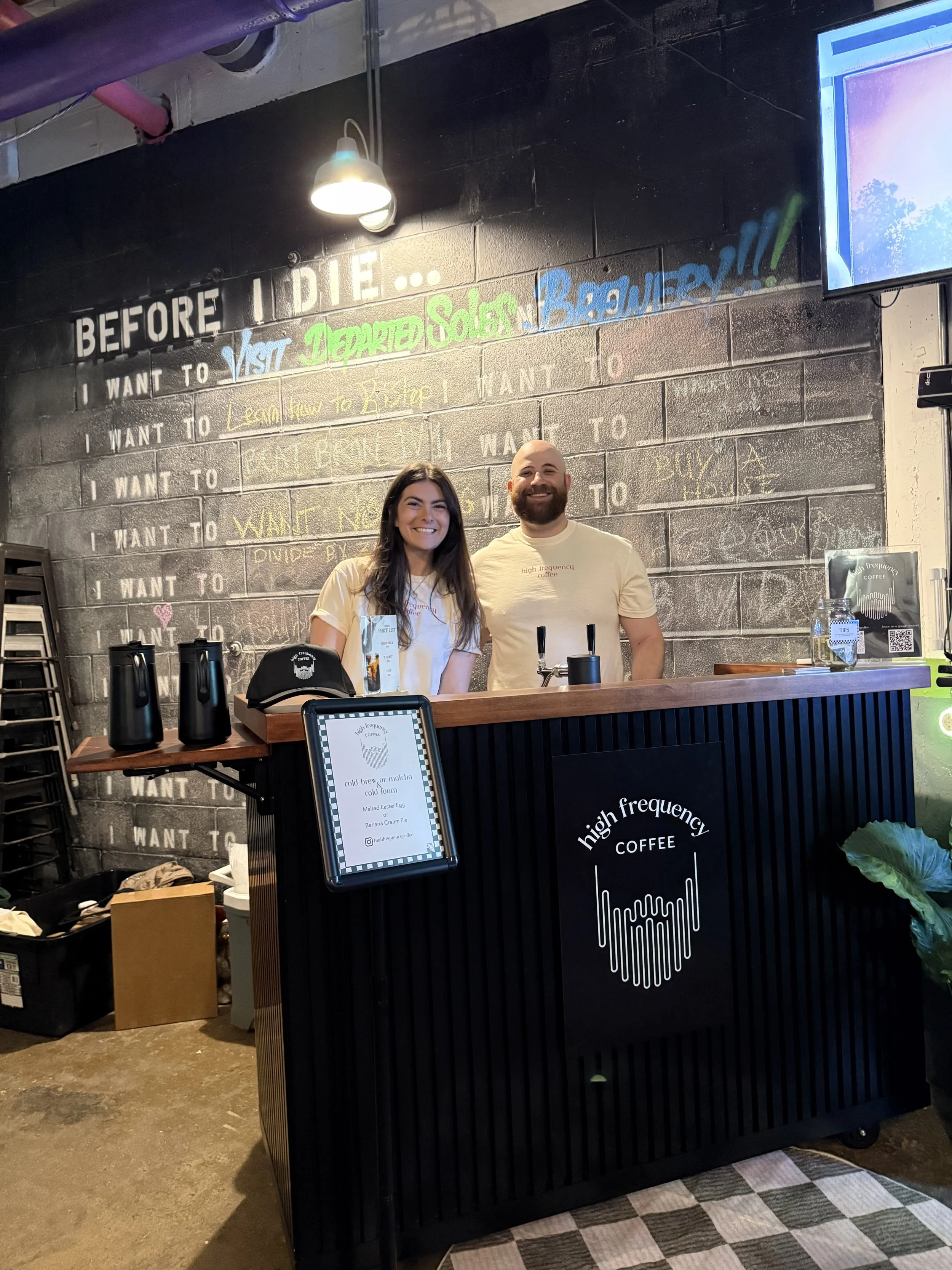 The founders of High Frequency Coffee are standing behind a coffee catering cart, with a tap for cold brew. The sign shows a menu for cold brew, matcha, and custom cold foams.