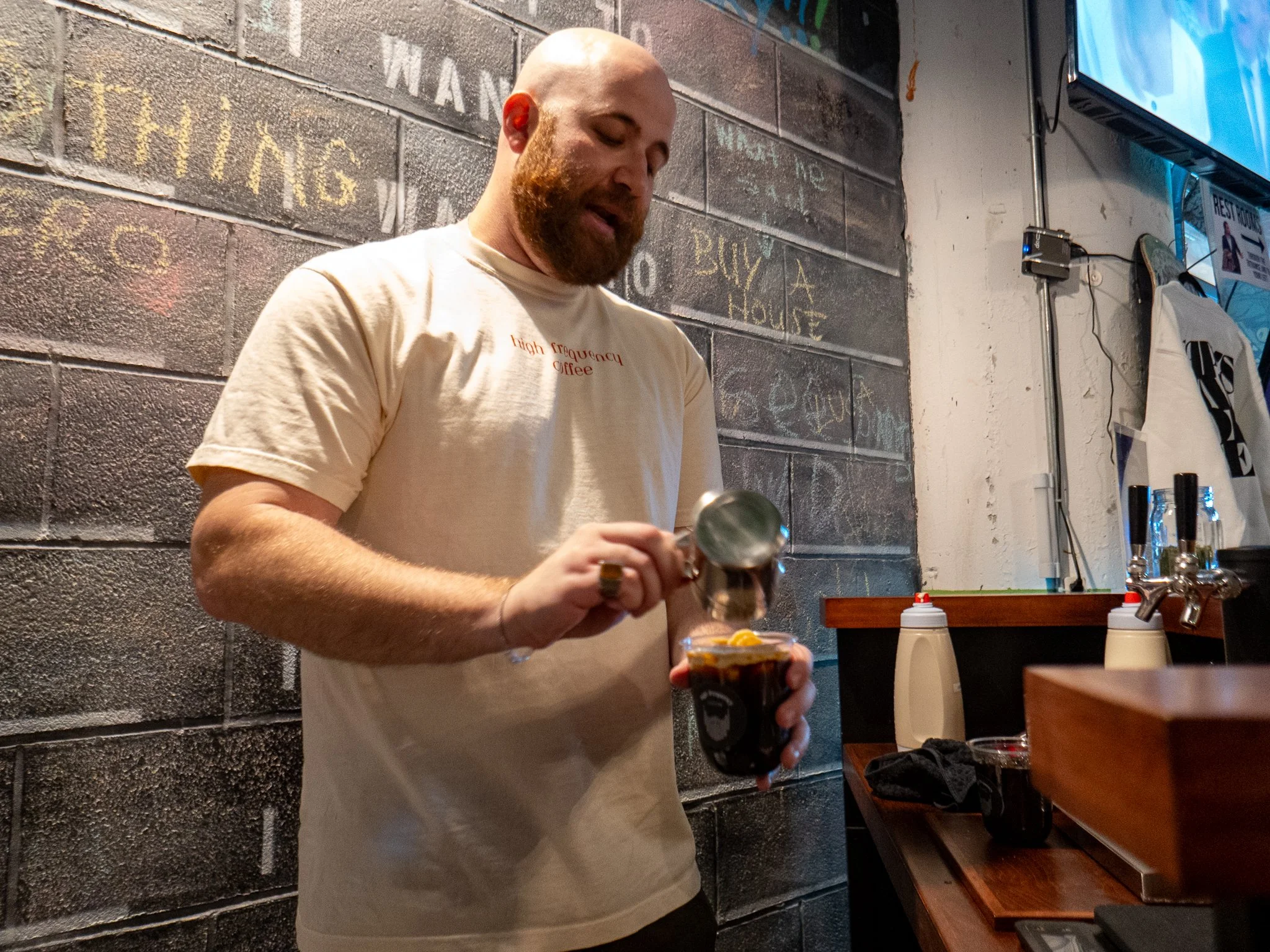 A man with a beard and a light-colored t-shirt is preparing an iced coffee with banana chips at a cafe counter.