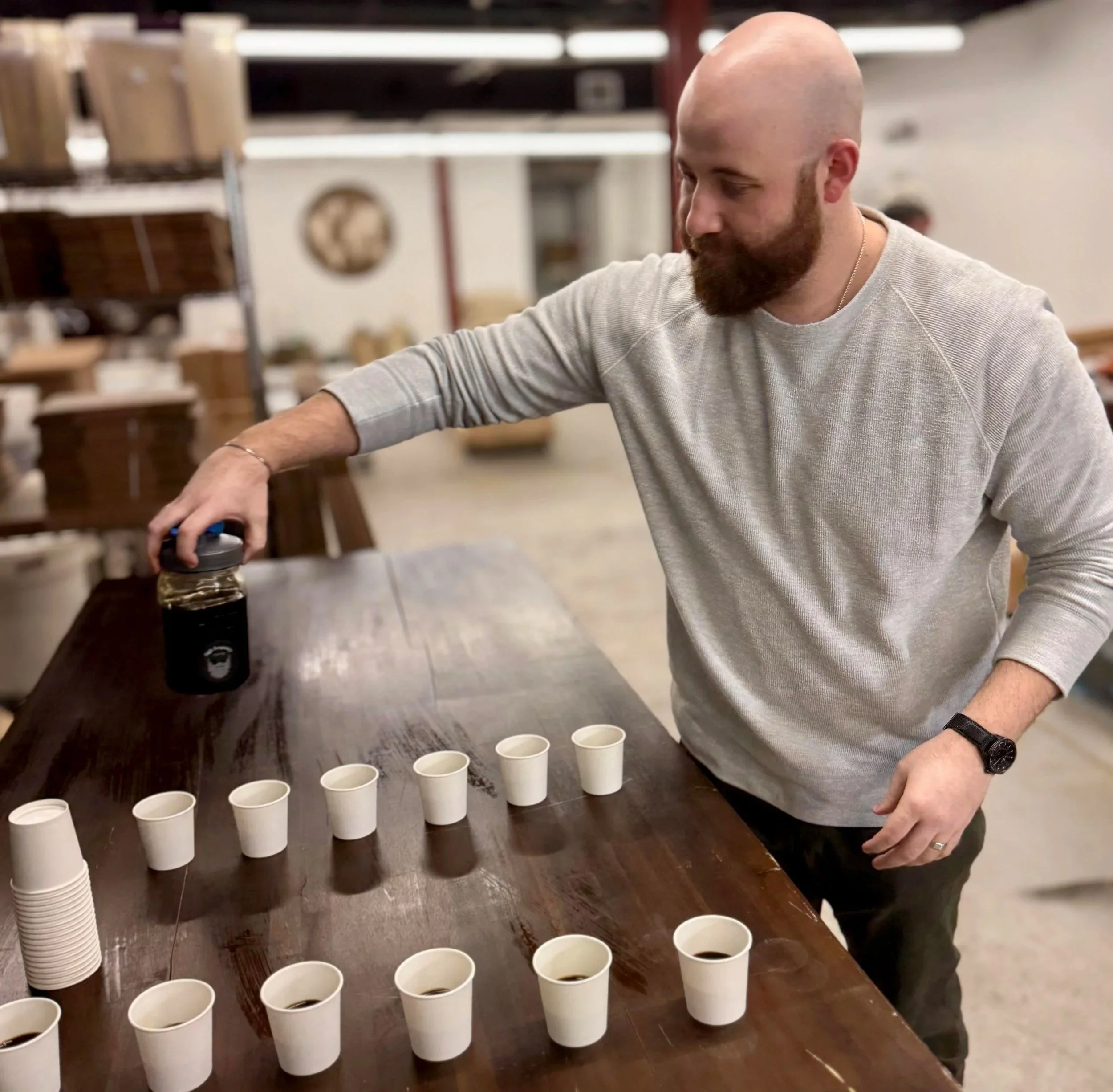 A man with a beard, wearing a gray long-sleeve shirt, is pouring coffee from a thermos into small white cups arranged on a wooden table in a warehouse or coffee shop setting.