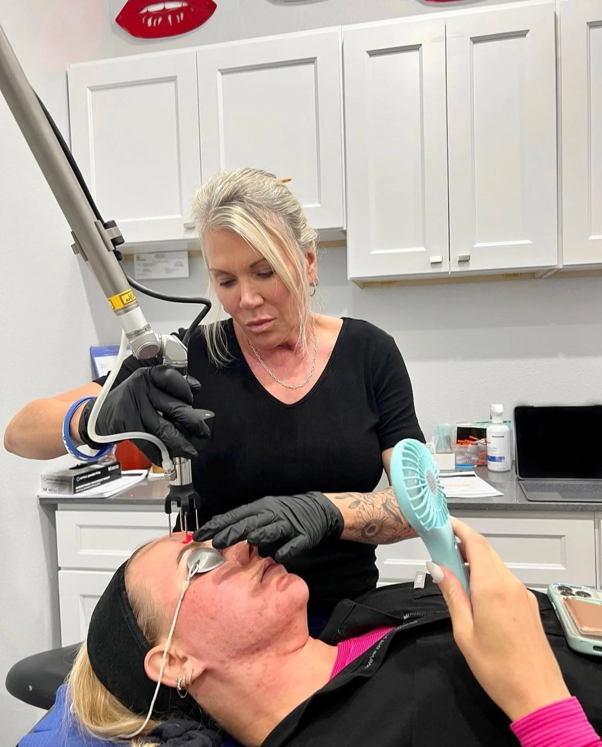A woman receiving a cosmetic laser treatment on her face from a professional in a clinic setting, with protective eyewear and a handheld fan for comfort.