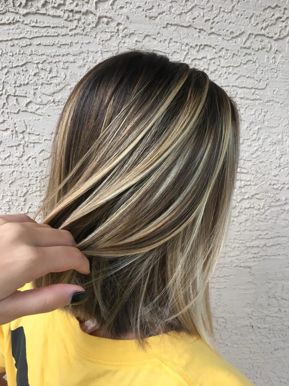 Person with shoulder-length, highlighted brown and blonde hair against a textured wall.