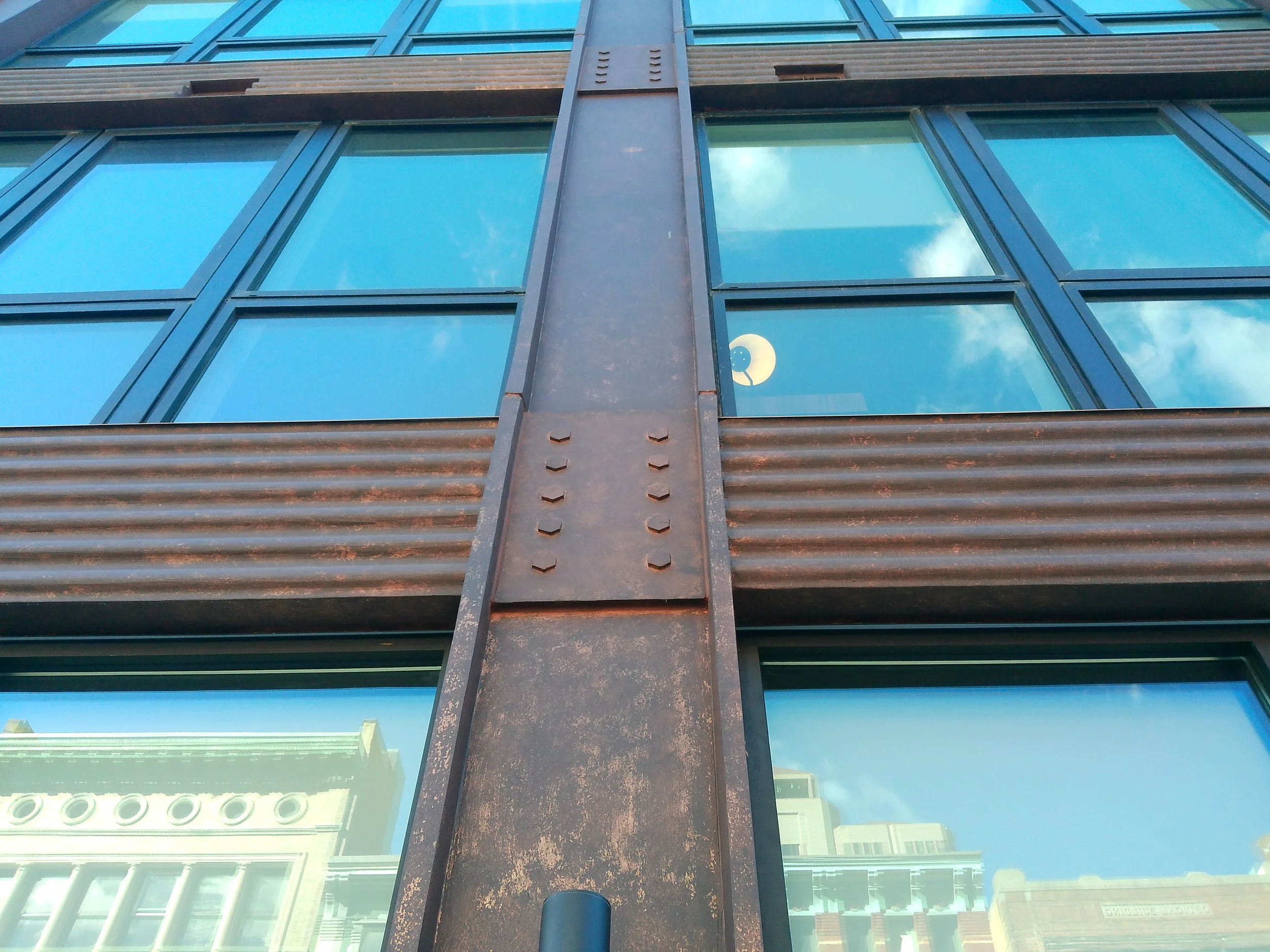Close-up view of a building facade with large glass windows and rusted metal panels, reflecting the sky and nearby structures.