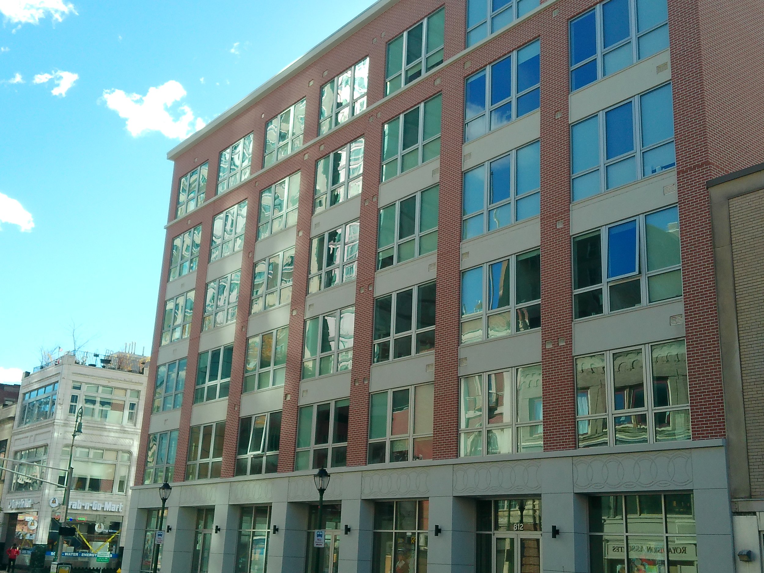 A multi-story modern building with a brick facade and large glass windows reflecting the sky and neighboring structures.