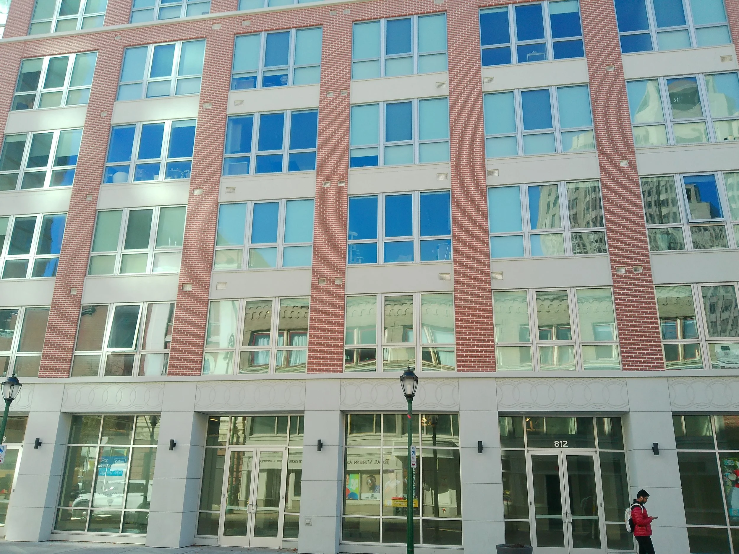 A multi-story modern building with a brick and glass exterior, with a storefront at the ground level and a person walking by on the sidewalk.