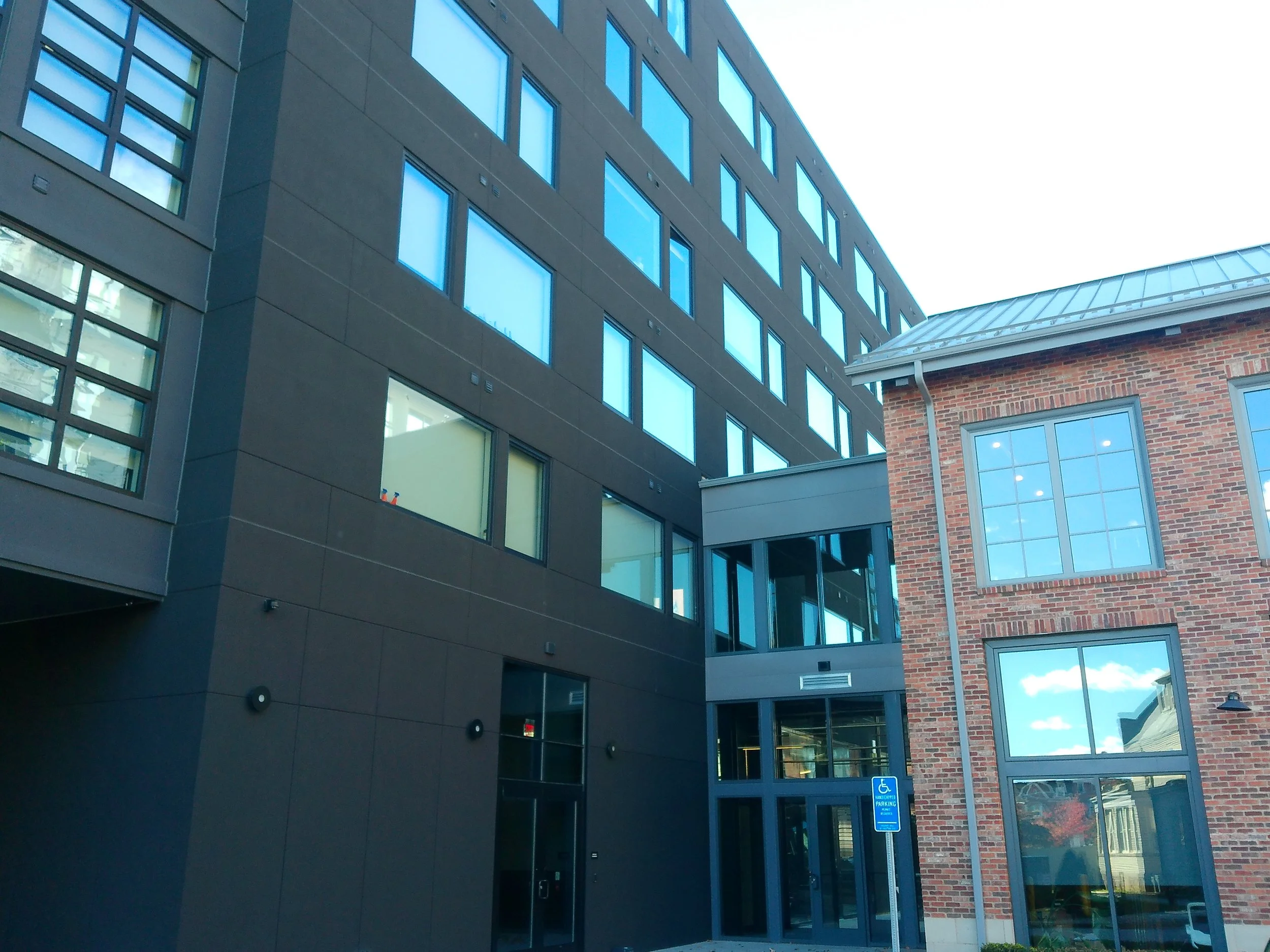 A modern multi-story building with large blue-tinted windows and a brick building with similar windows, and a parking sign for handicap parking