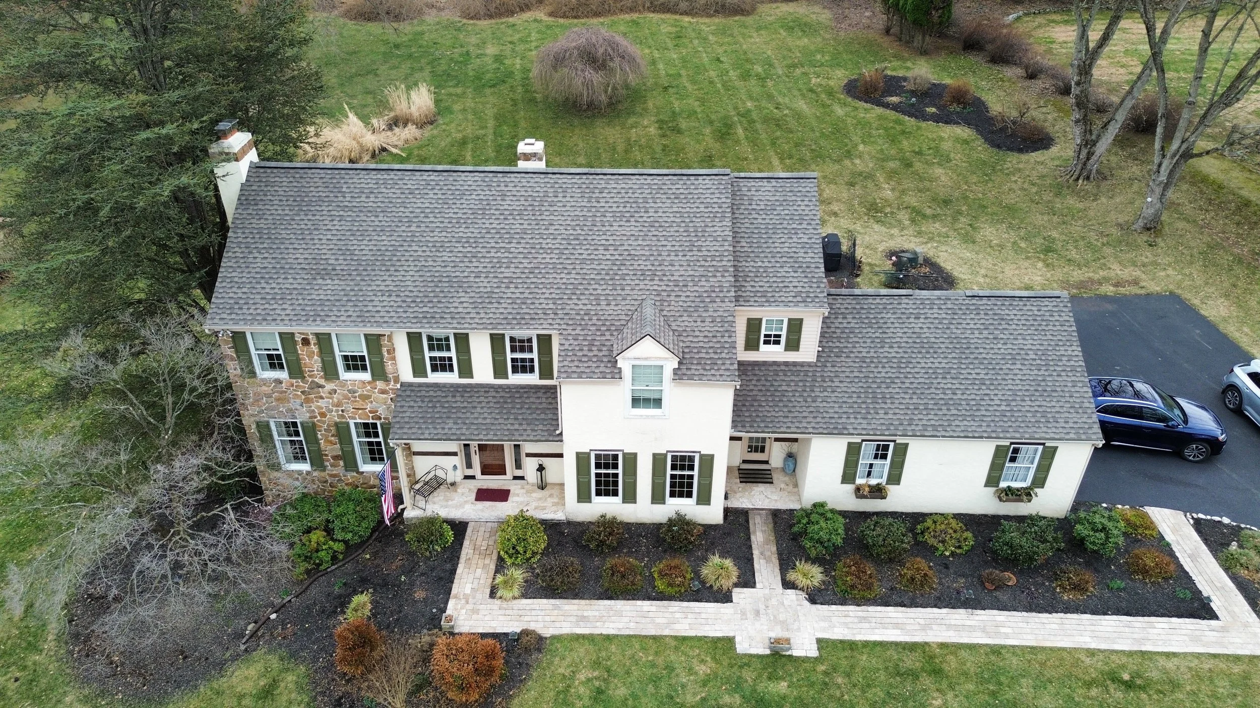 Aerial view of a two-story house with a stone and siding exterior, green shutters, a front porch with steps, a black asphalt driveway with two cars, and a landscaped yard with bushes, trees, and a lawn in West Chester, PA.