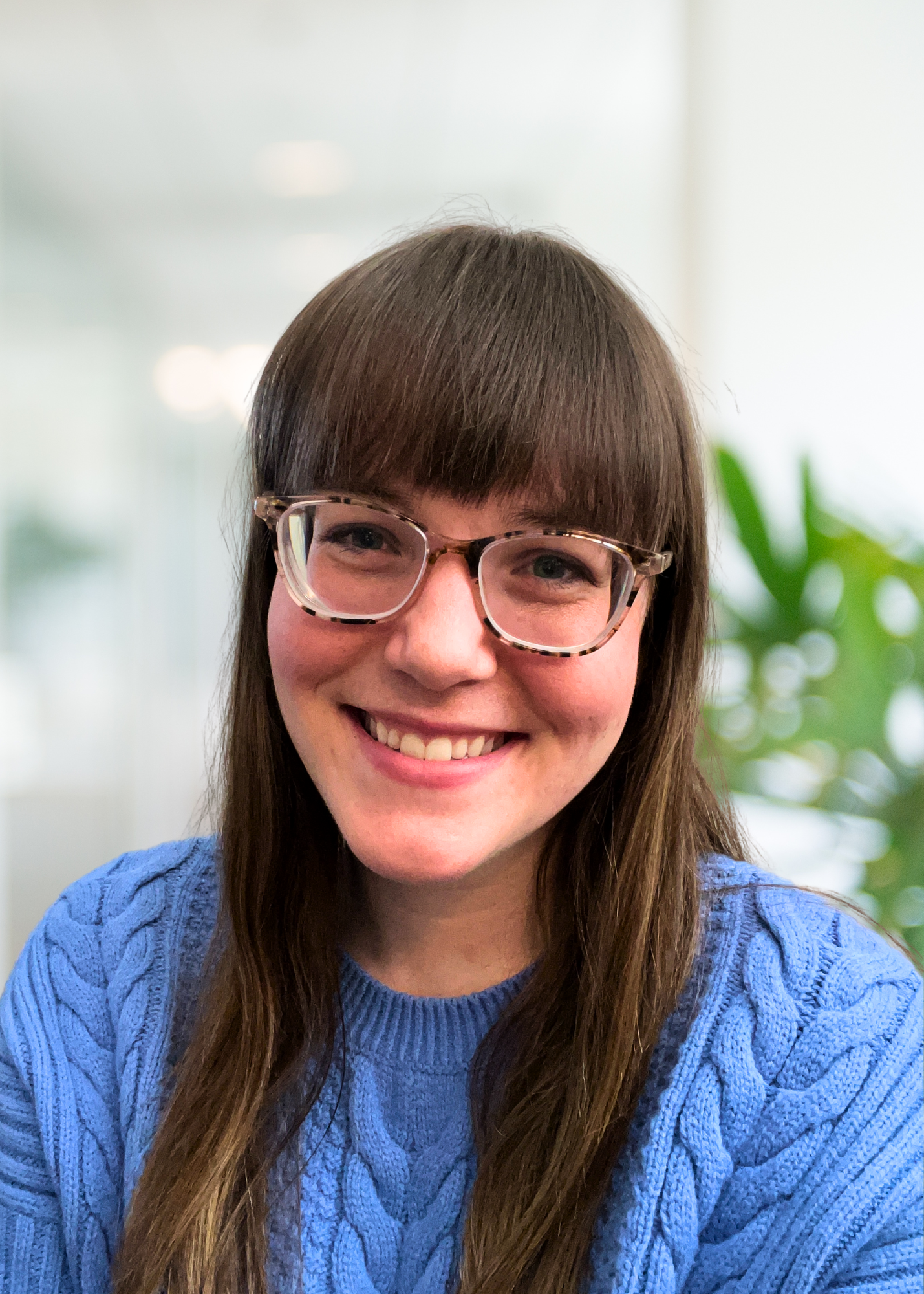 Close-up of a young woman with long brown hair, blue eyes, and a light smile, wearing a blue textured top, indoors with a blurred background and greenery.