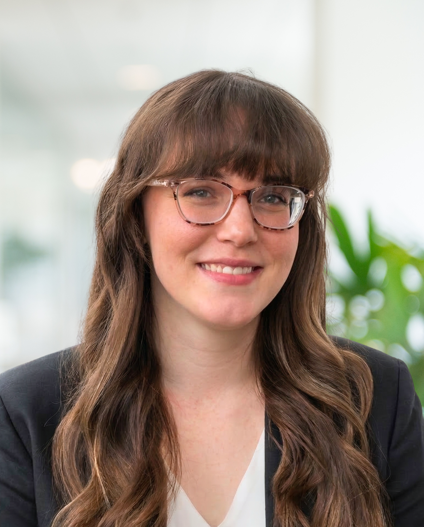 A young woman with long brown hair, blue eyes, and a subtle smile, wearing a blue top, standing indoors with a blurred background of windows and a green plant.