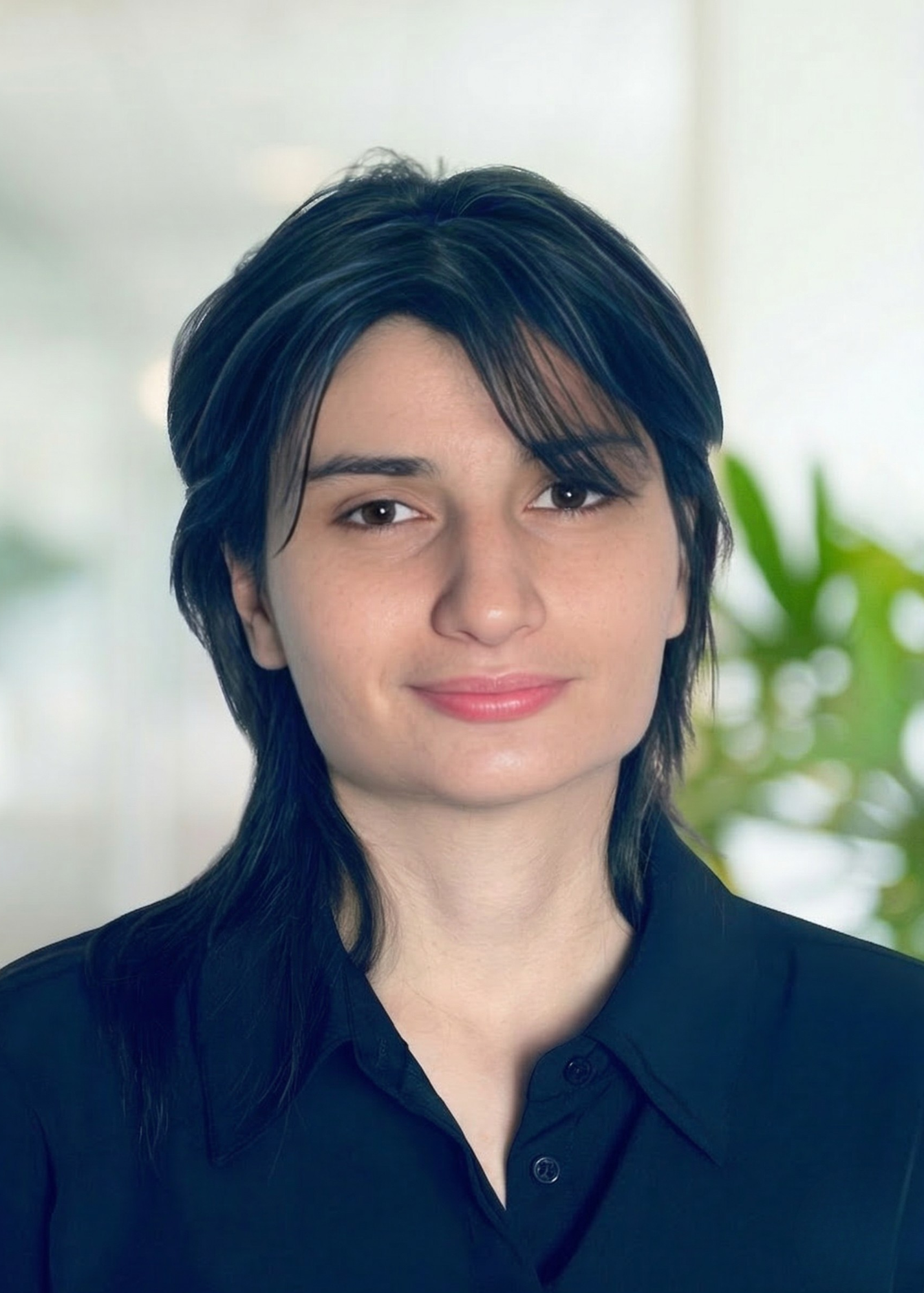 A young adult with short, dark hair and fair skin, wearing a black collared shirt, smiling softly, with a blurred indoor background and a green plant.