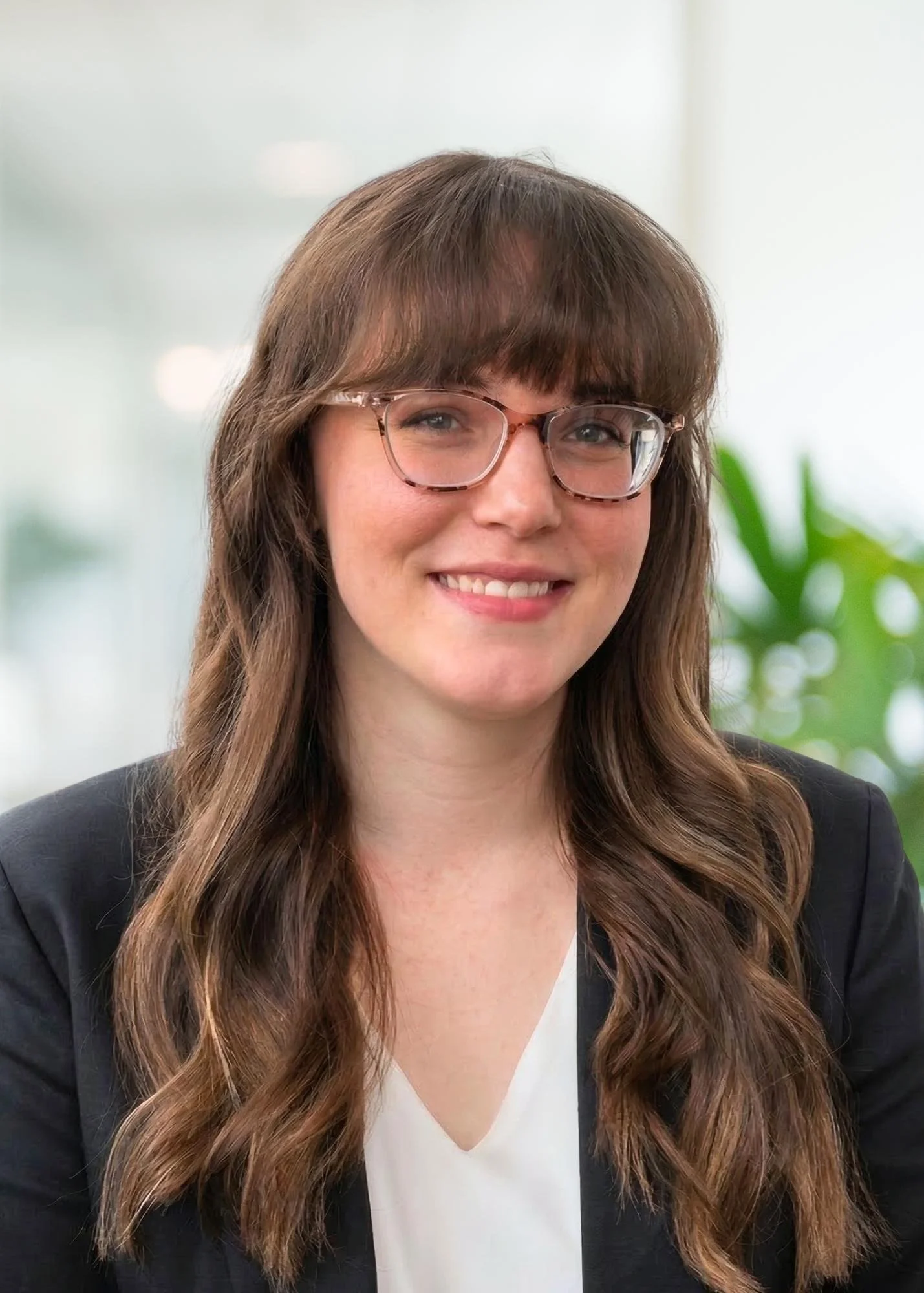 Close-up of a young woman with long brown hair, blue eyes, and a light smile, wearing a blue textured top, indoors with a blurred background and greenery.
