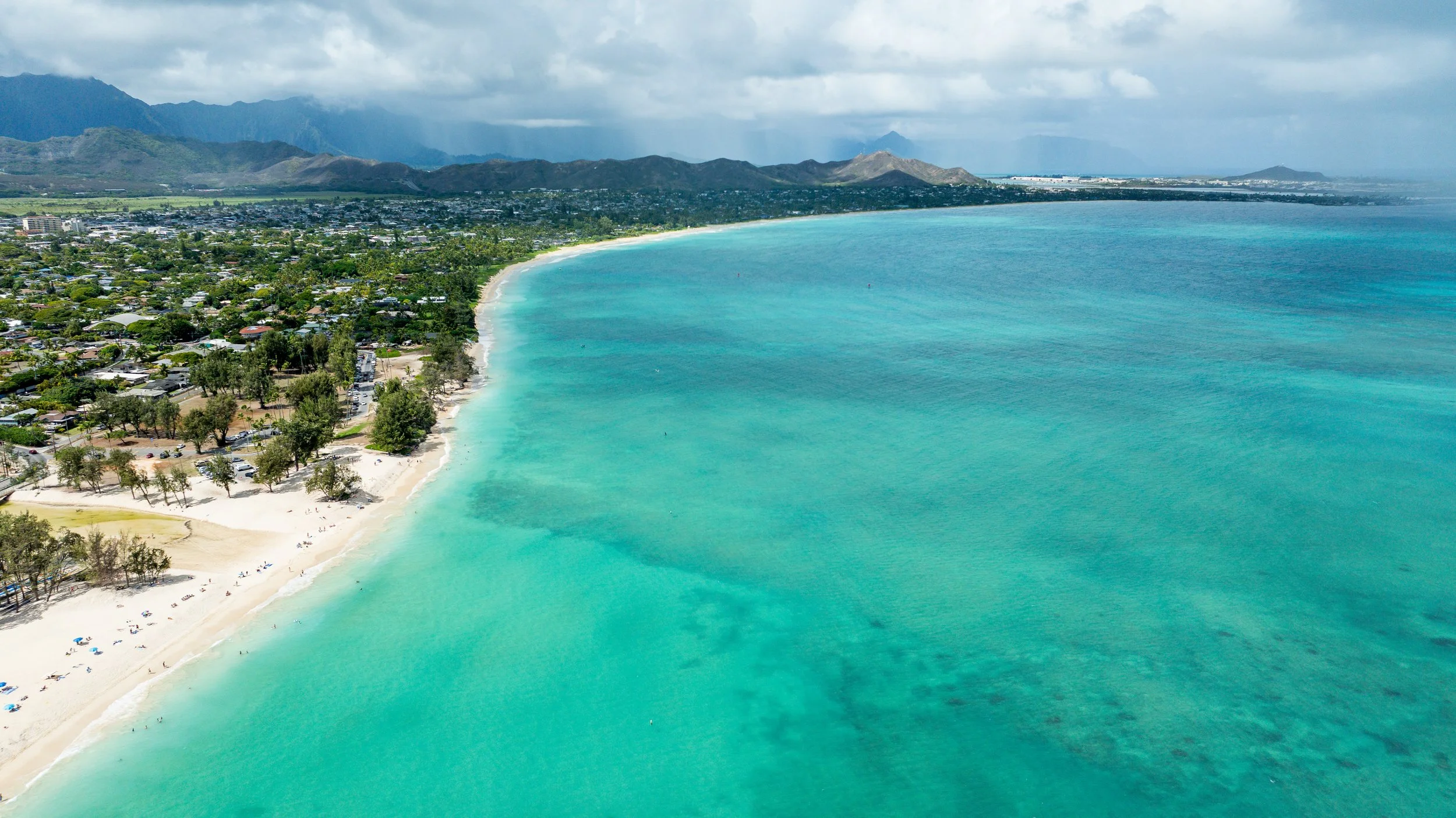 An aerial view of a beach with turquoise waters, a shoreline with trees and parked cars, and a town with buildings in the background, with mountains behind.