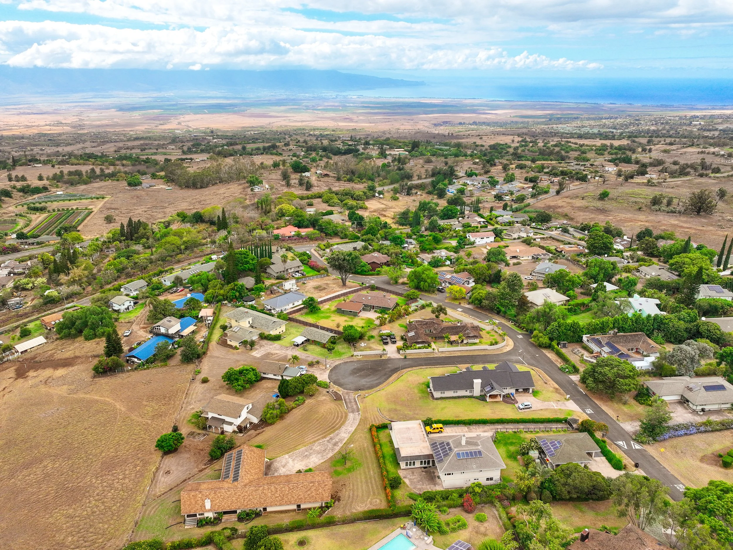 Aerial view of a residential neighborhood with houses, trees, and green lawns, set against a landscape with fields and mountains in the distance, under a partly cloudy sky.