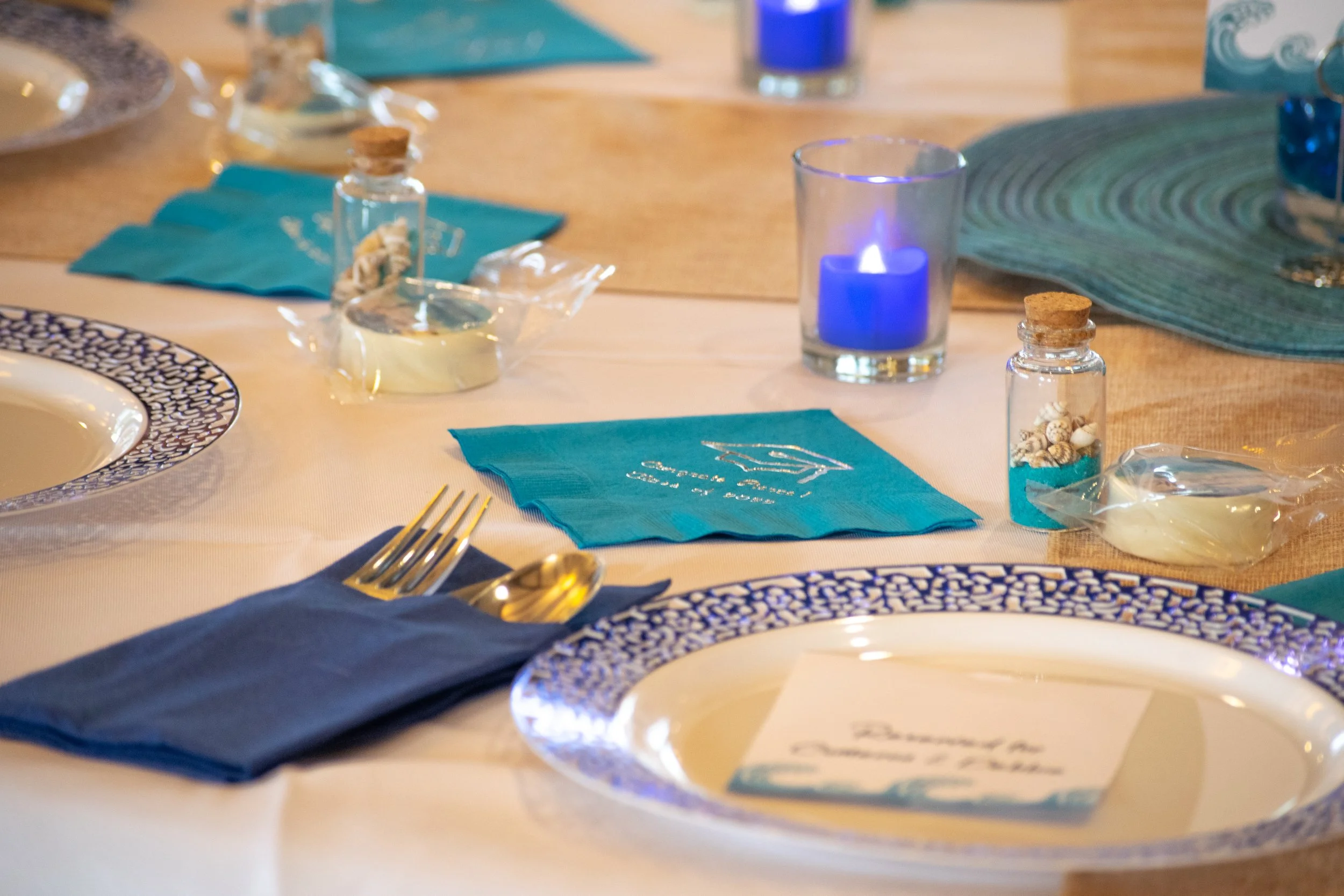 A decorated dining table set with blue and white plates, gold utensils on a blue napkin, teal napkins with writing, a blue candle in a glass holder, small bottles filled with seashells, and a place card.