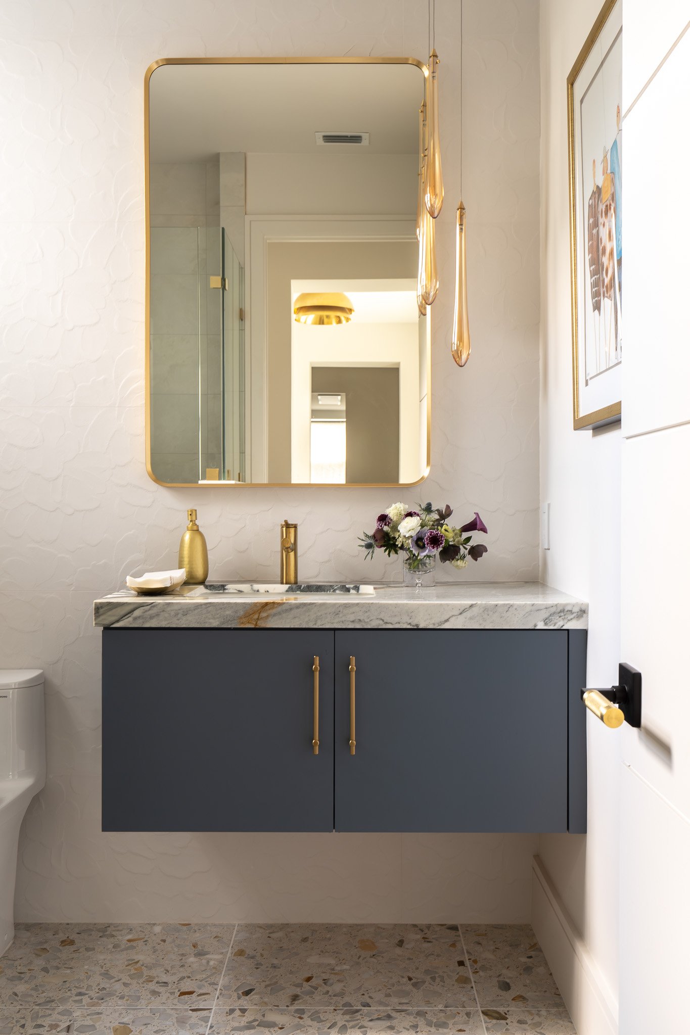 A bathroom with a large mirror framed in gold, a navy blue cabinet with gold handles, a marble countertop with a gold soap dispenser and vase of flowers, and a wall-mounted black and gold toilet paper holder. There are hanging glass pendant lights an