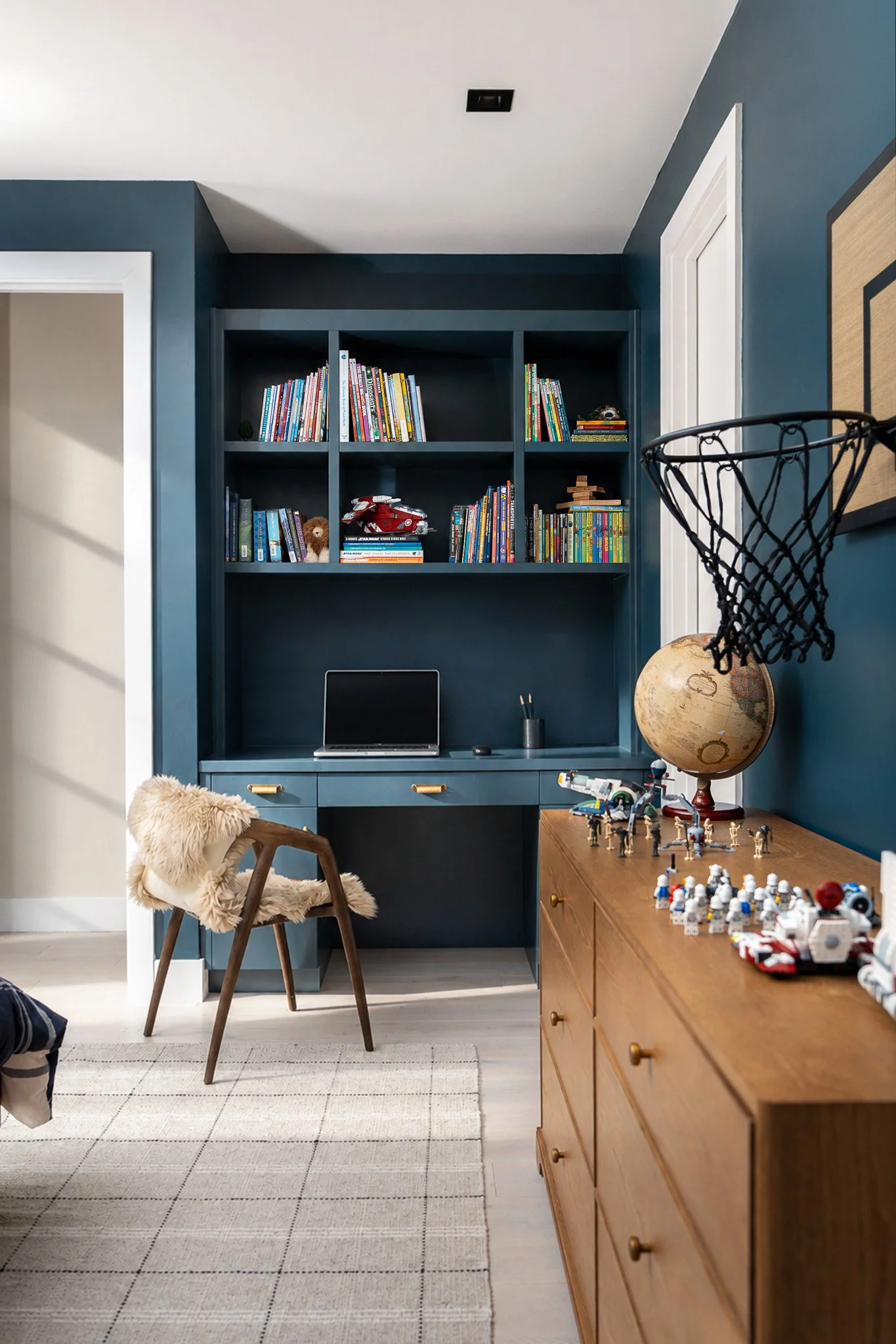 A room with a blue wall and a built-in bookshelf filled with colorful books and toys, including a toy car and a small stuffed animal. There is a wooden dresser with a globe and toy figures on top. A chair with a fur blanket is placed in front of the 