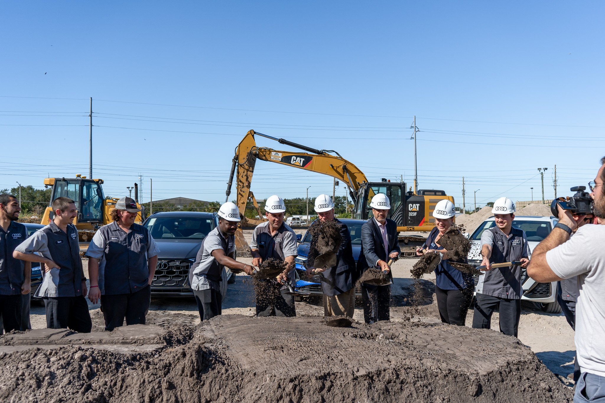 Group of people attending a groundbreaking ceremony, some digging into the dirt with shovels, with construction equipment and cars in the background, and a clear blue sky overhead.