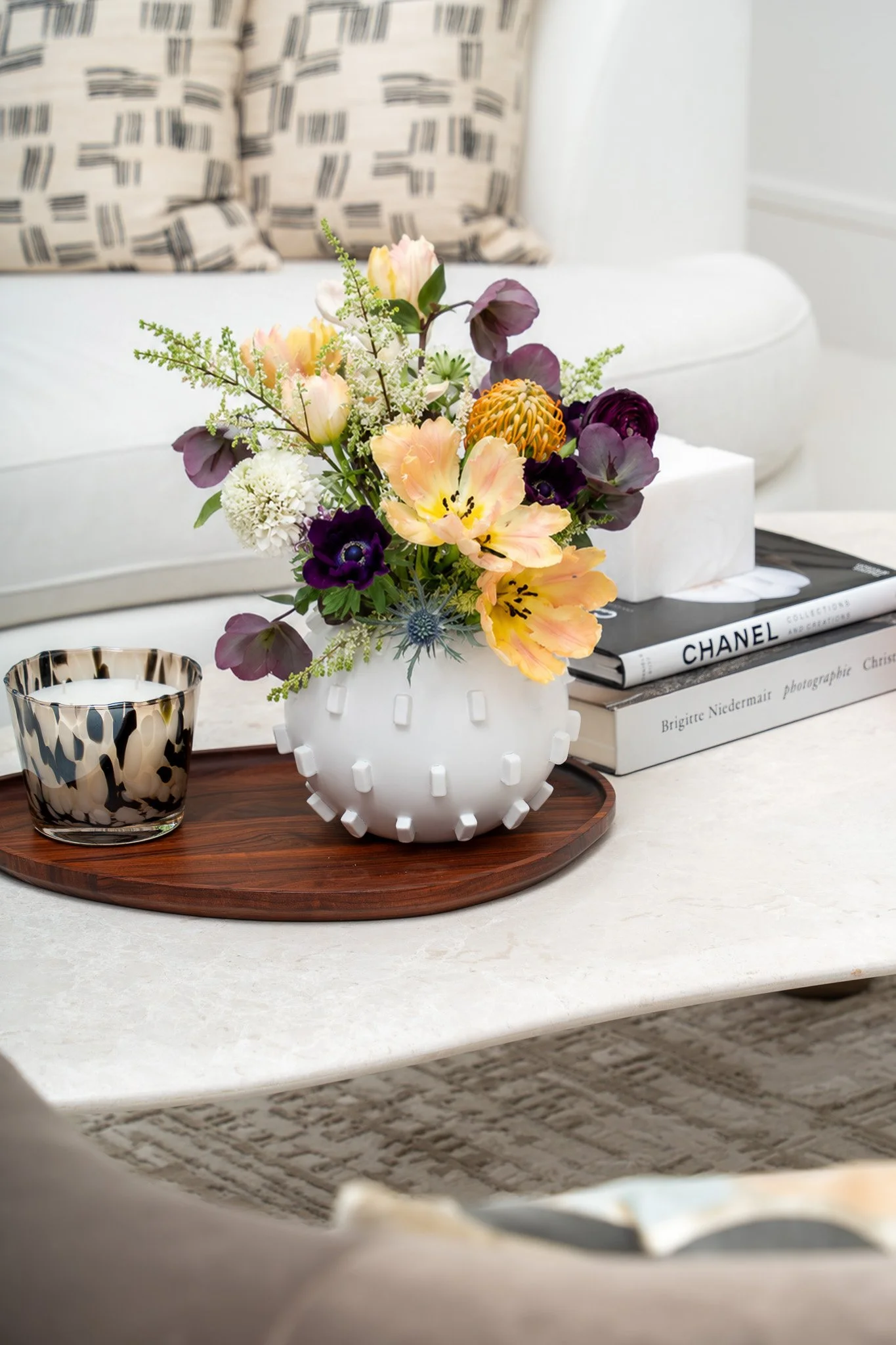 A bouquet of colorful flowers in a white vase on a round wooden tray, placed on a white table. A black and white patterned candle holder is nearby. In the background, a white couch with a black and white patterned pillow and a stack of fashion books,