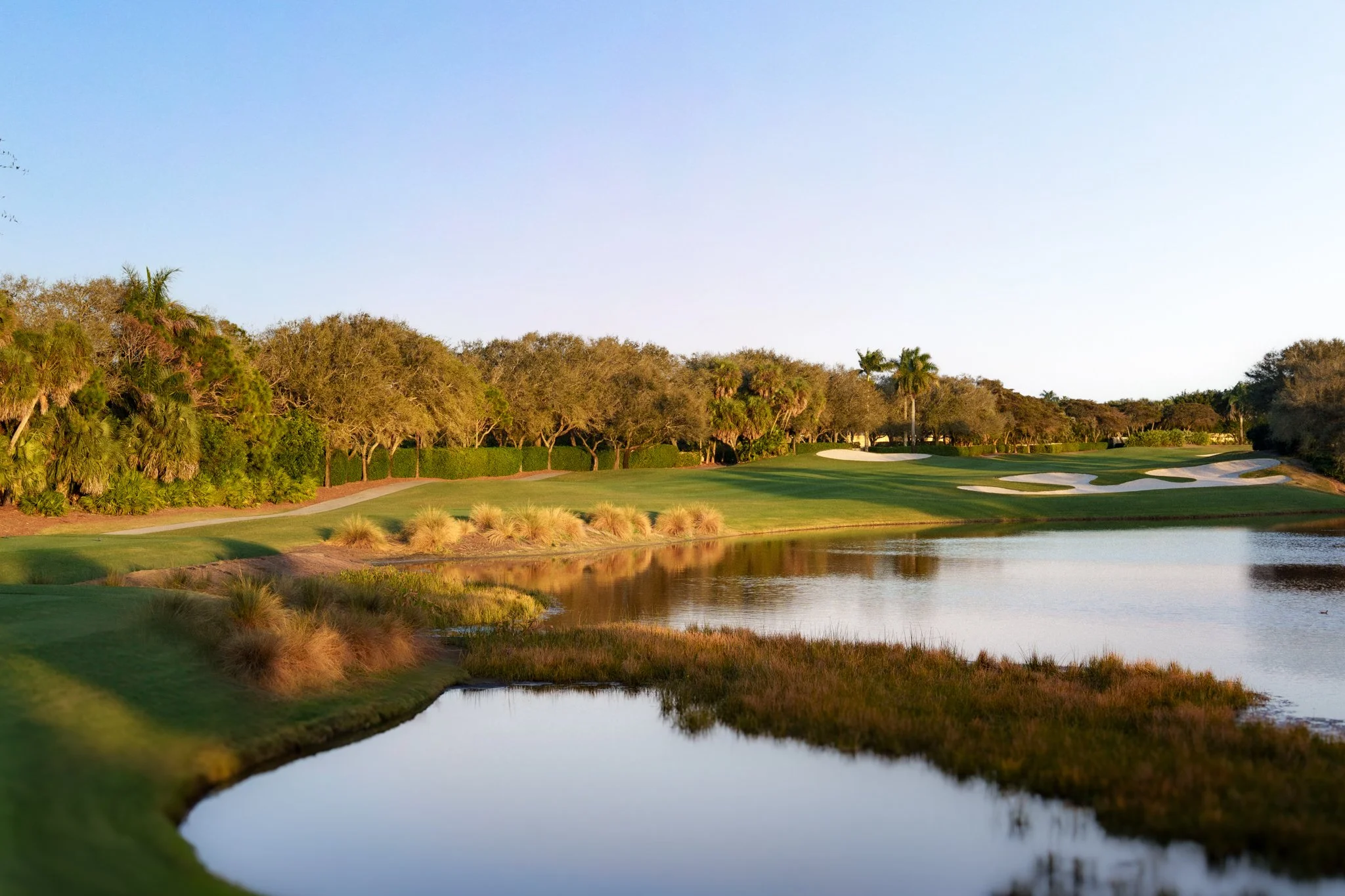 A scenic view of a golf course near a water hazard with trees and sand traps, under a clear sky.