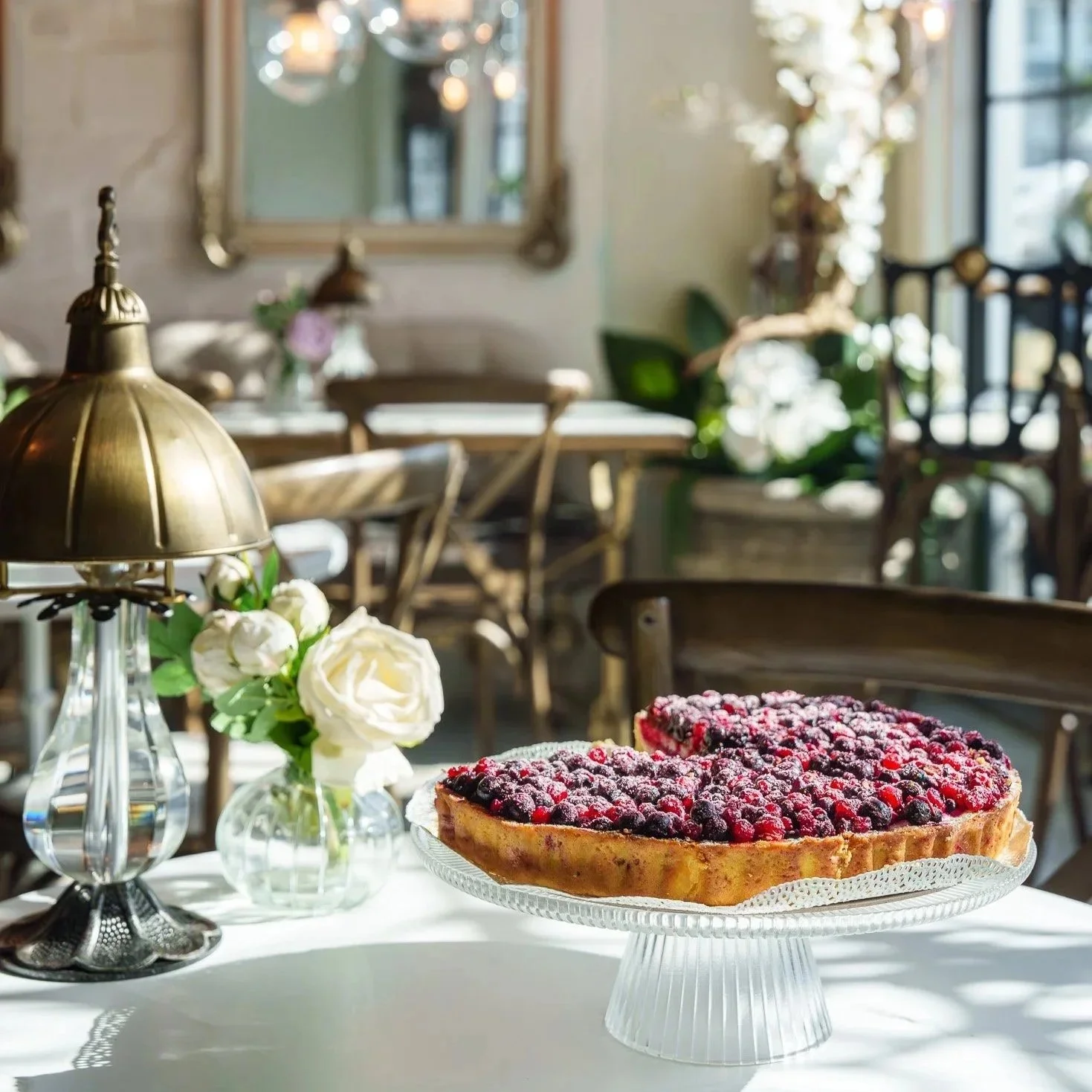 A fruit tart topped with mixed berries on a glass cake stand, a white flower in a glass vase, a vintage brass table lamp, and a dining room with chairs, a mirror, and hanging lights.