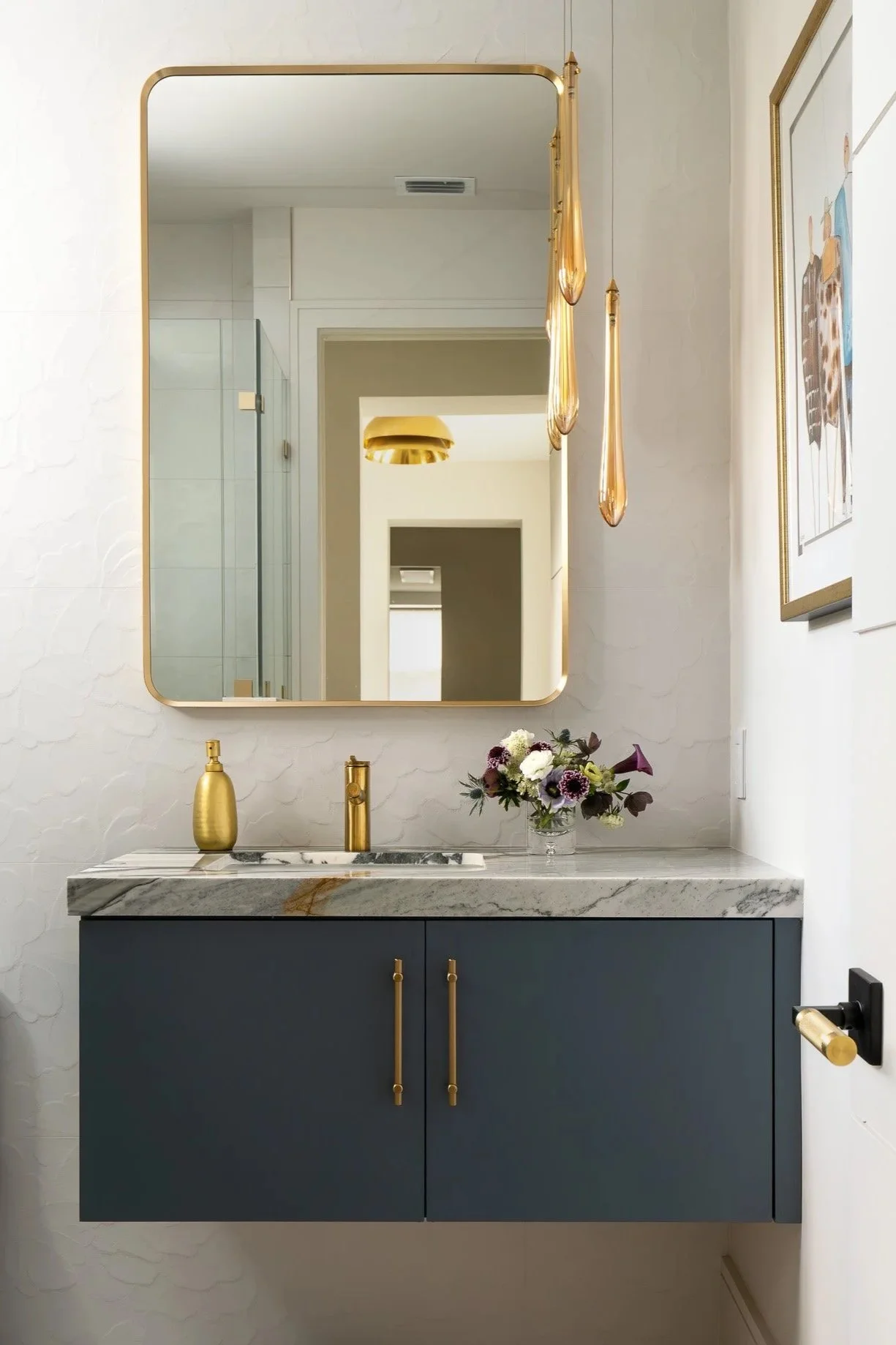 A modern bathroom vanity with a marble countertop, dark blue cabinets with gold handles, a gold soap dispenser, a gold faucet, a vase with flowers, a large framed mirror, floating gold pendant lights, and framed artwork on the wall.