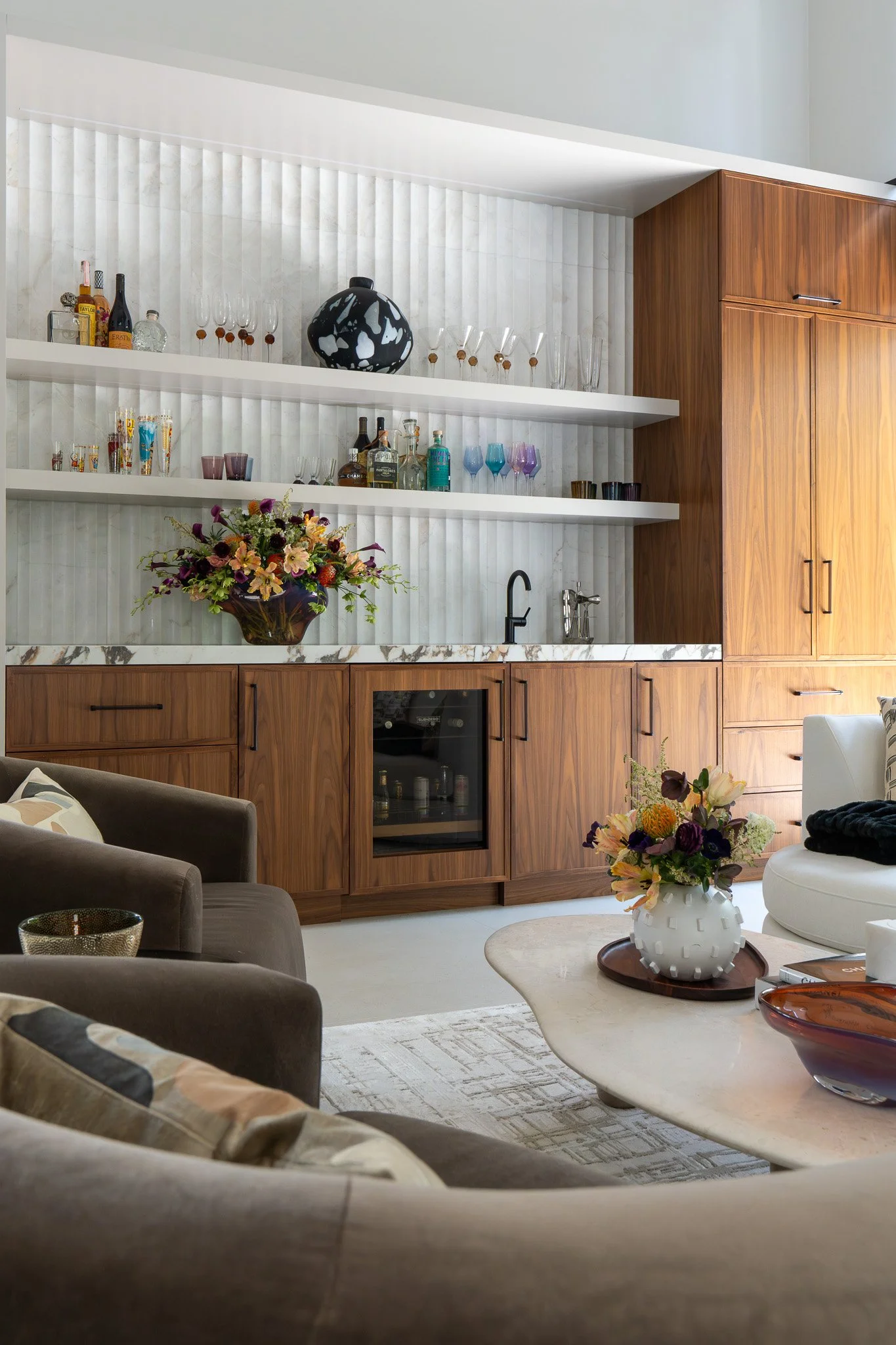 Living room with beige sofas, a white marble coffee table with decorative vases, and a book. In the background, wood cabinetry and open shelves with glasses and bottles. Floral arrangements decorate the space.