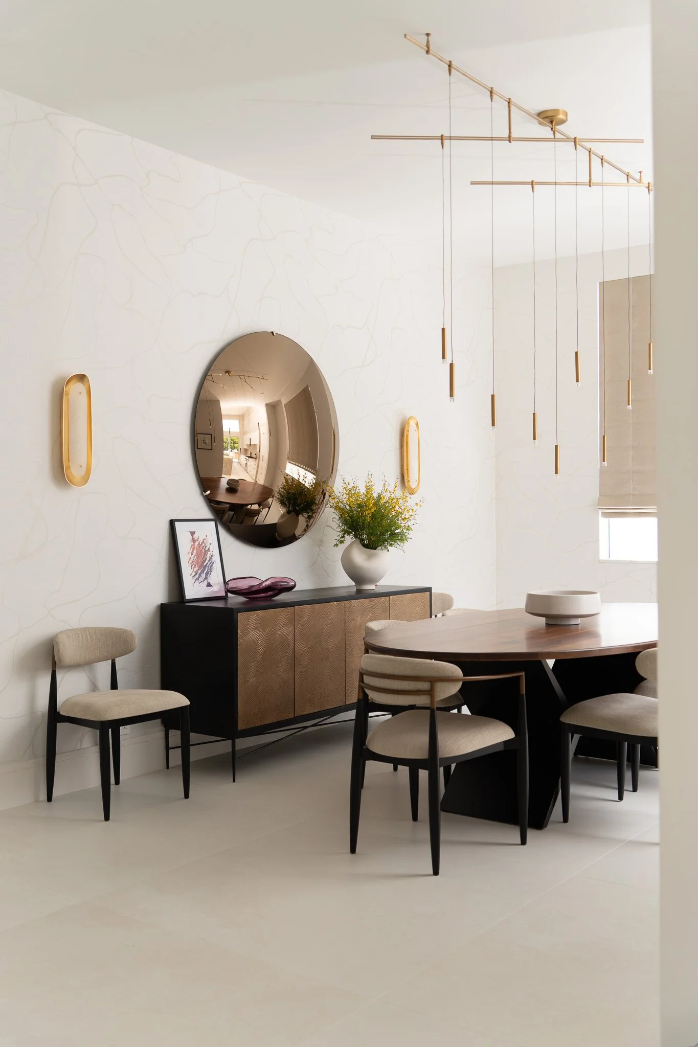 Modern dining room with a round wooden table, six beige chairs with black legs, a black sideboard with gold details, a white vase with flowers, a framed artwork, a round mirror, and decorative lamps on the wall. Gold pendant lights hang from the ceil