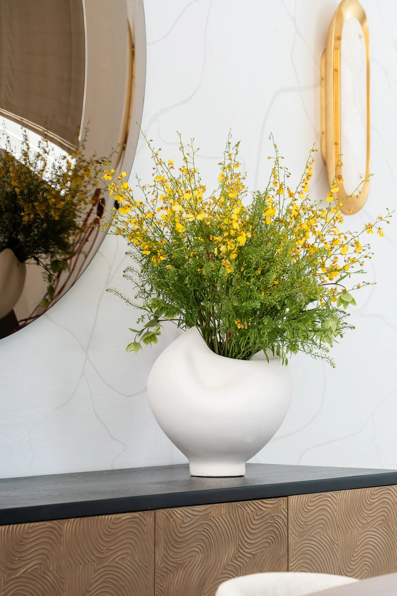 A white vase with a round body and narrow neck holding a bouquet of yellow flowers and green foliage, placed on a black surface. A mirror and a wooden shelf are visible on the wall behind the vase.