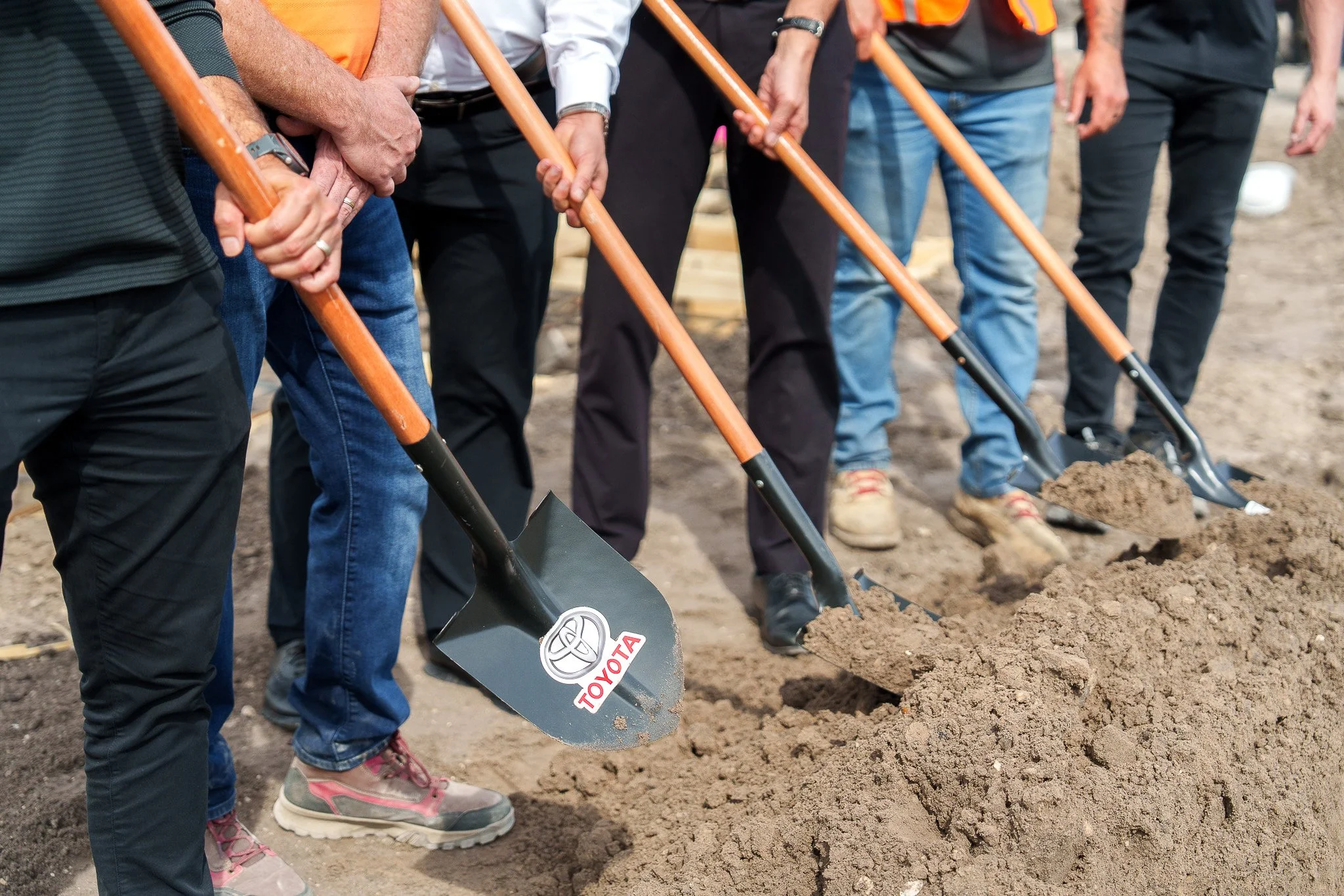 Multiple people standing in a row holding shovels with the blades in the dirt, participating in a groundbreaking ceremony. The shovels have the Toyota logo on them, indicating a corporate-sponsored event.