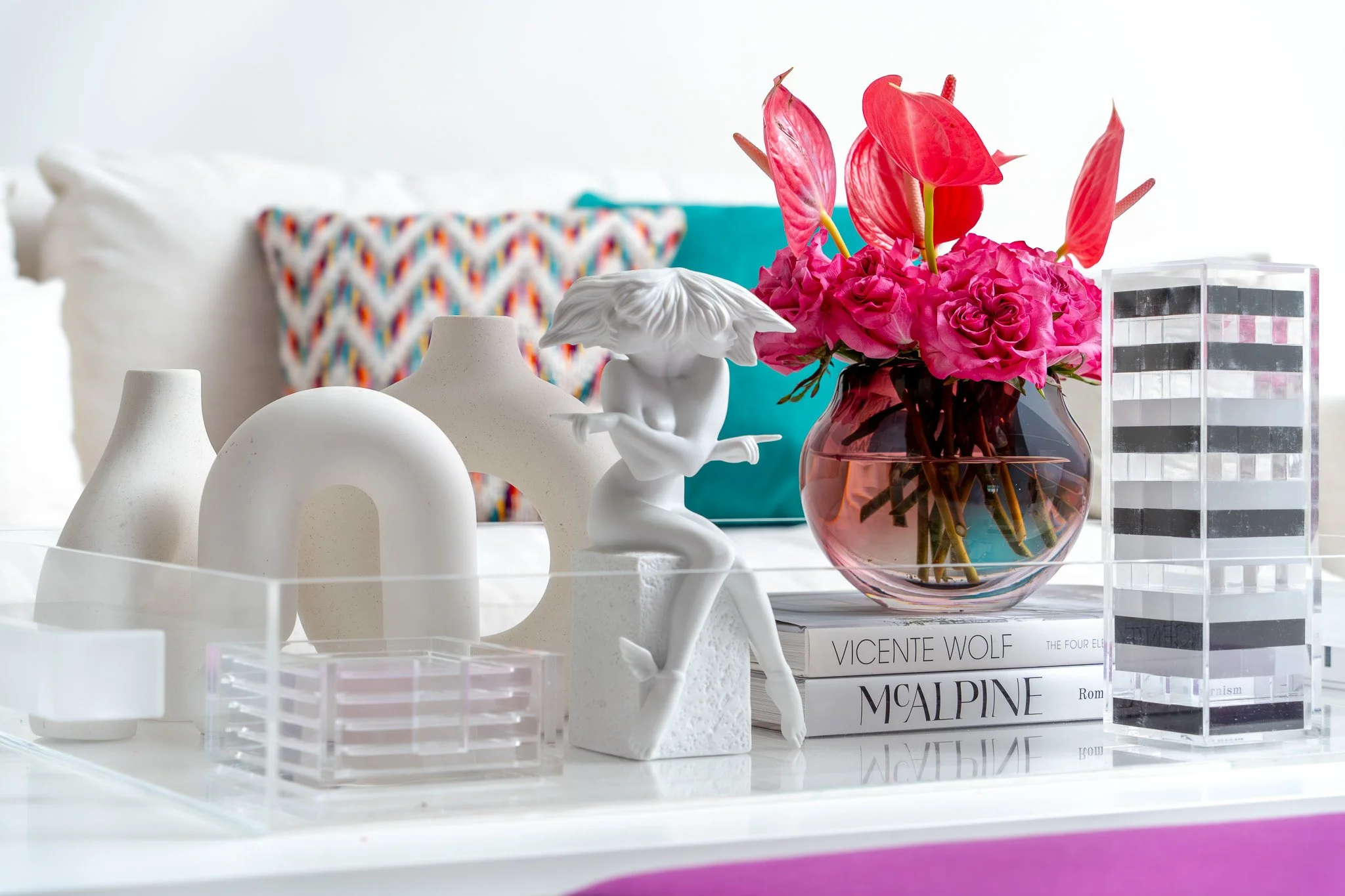 A collection of decorative objects including white ceramic vases, a small white artistic sculpture of a child, and a glass vase with a bright pink floral arrangement of anthuriums and roses. Background features colorful geometric pillows and stacked 