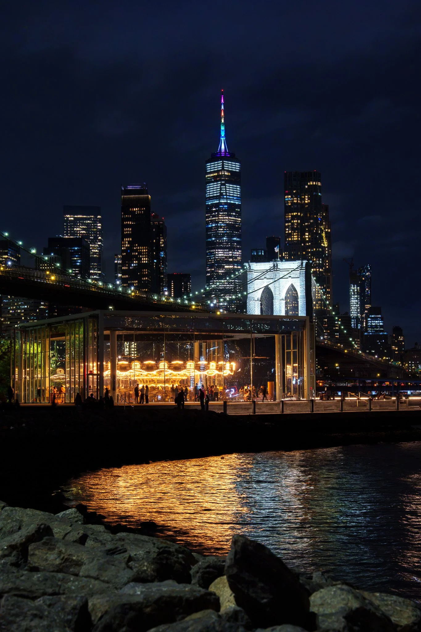 Night view of the New York City skyline featuring the Empire State Building illuminated with colorful lights, reflected in the water below, with a promenade and people sitting and walking along the waterfront.