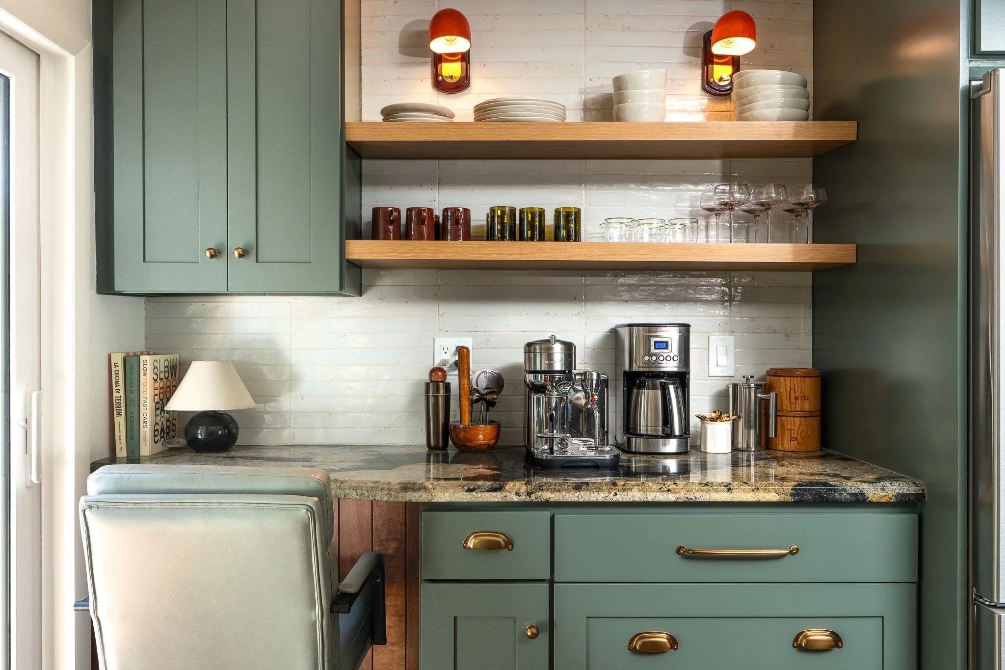 A kitchen with a granite countertop, green cabinets, open wooden shelves with plates, bowls, glasses, and cups, a coffee maker, a toaster, and various kitchen accessories, with a white brick wall and a window.