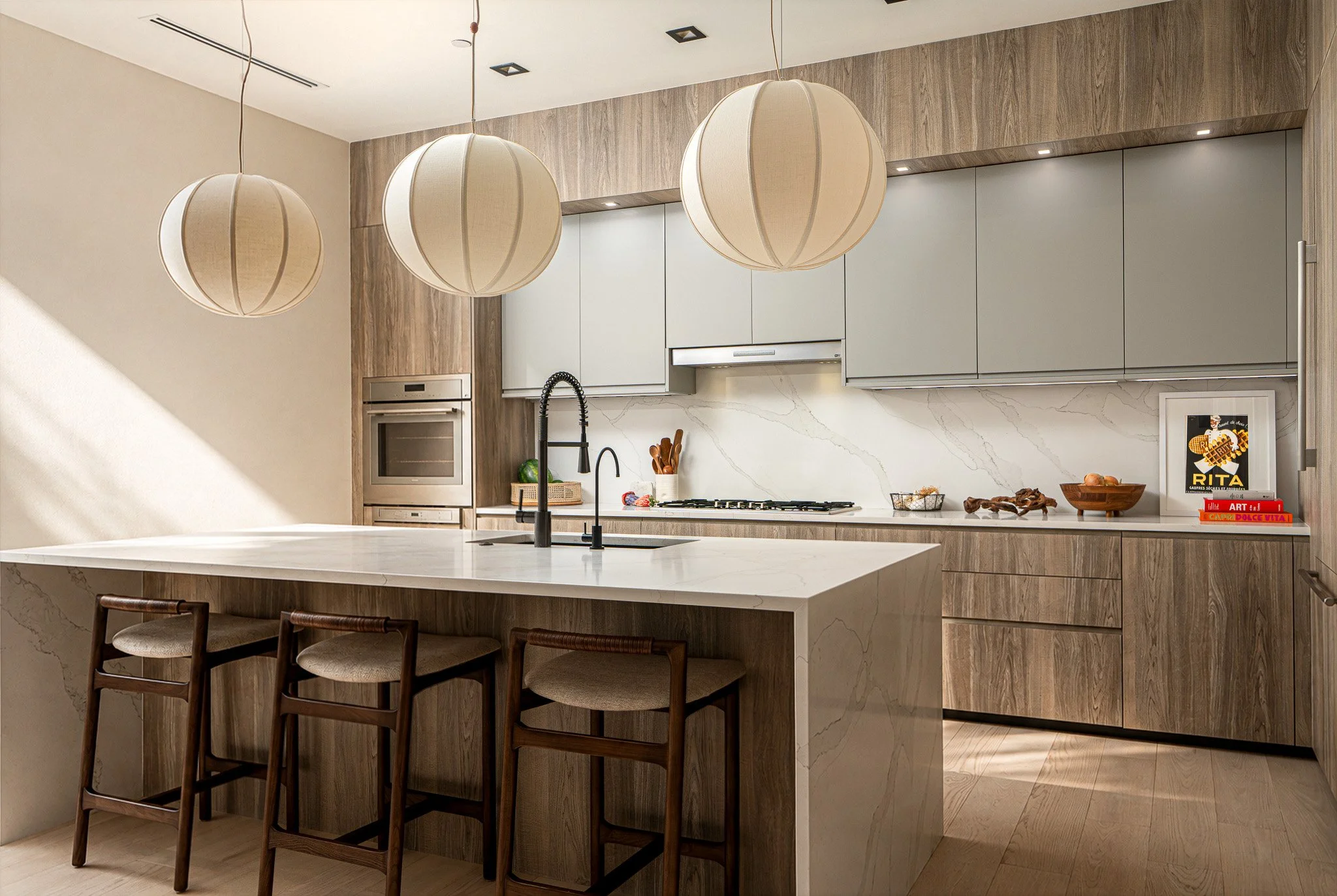 Modern kitchen with beige and wood cabinetry, marble backsplash, center island with white countertop, three beige pendant lights above, black faucet, and bar stools at the island.