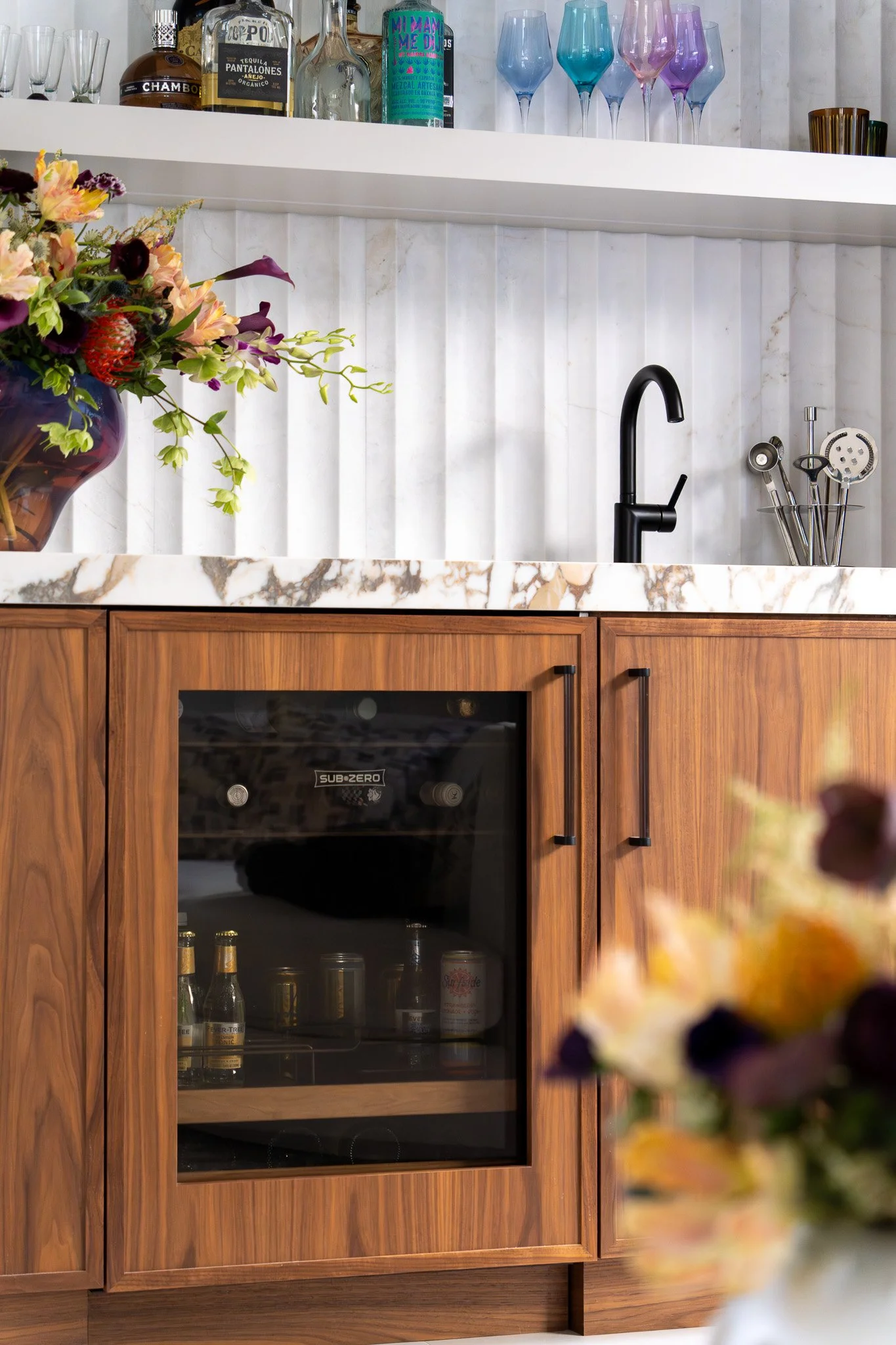 Kitchen with wooden cabinetry, marble countertop, black faucet, cocktail glasses, liquor bottles, and a flower vase.