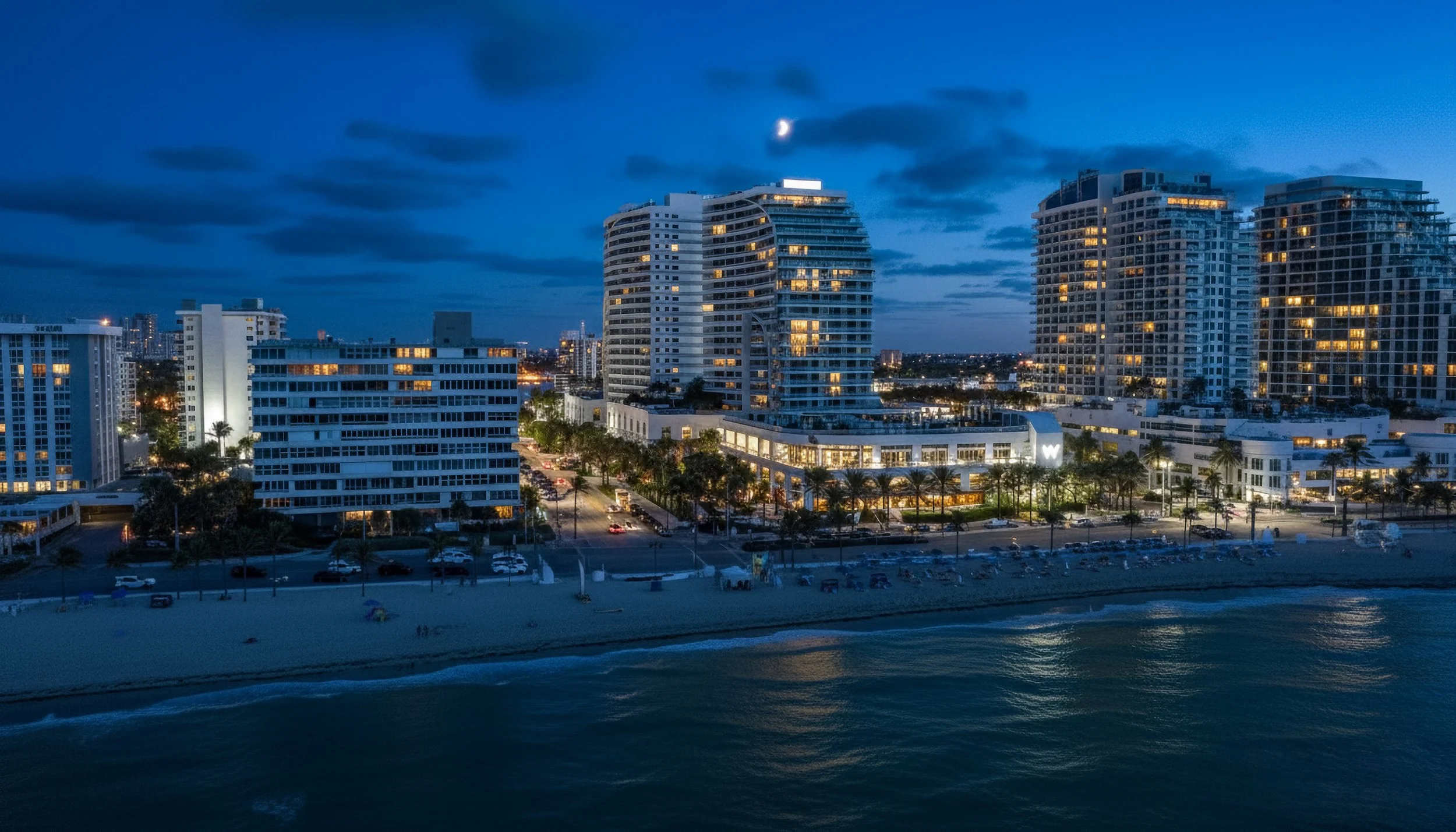 Nighttime cityscape with high-rise buildings along a beach, some windows lit, a partly cloudy sky, and the moon visible.