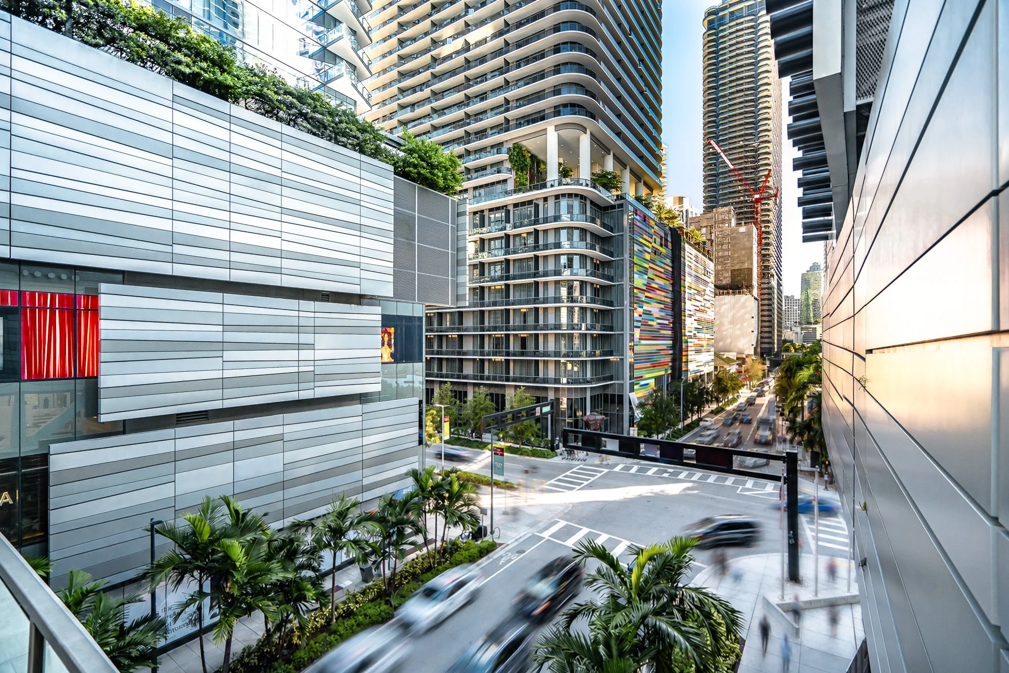 Modern cityscape with tall buildings, including a high-rise with rounded balconies and greenery on the upper levels, a colorful mural on one building, and a busy street with moving cars, palm trees, and sidewalk cafes.