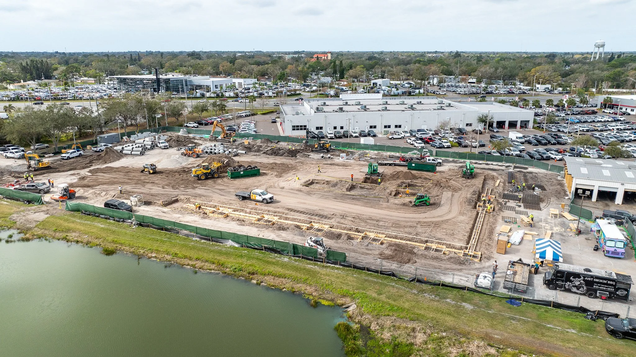 An aerial view of a construction site next to a body of water with multiple construction vehicles and workers, surrounded by parking lots and commercial buildings.
