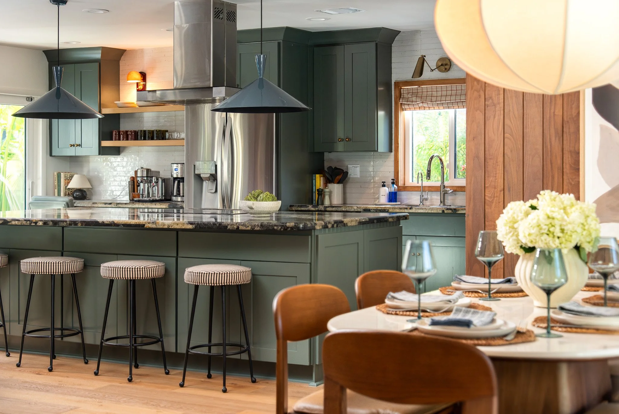 A modern kitchen with a large island counter, green cabinets, and stainless steel appliances. There are three striped bar stools in front of the island. The kitchen has natural light coming through a window and a dining area with a table set with win