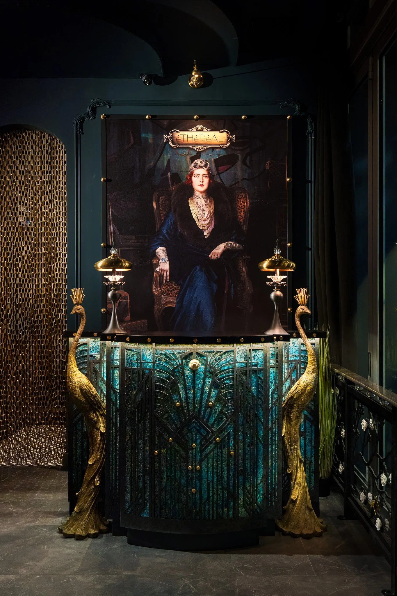 A decorative interior display featuring a large portrait of a woman with dark hair and tattoos, sitting on a throne-like chair. The display includes gold peacock sculptures, two gold lamps, and intricate black and gold architectural elements.
