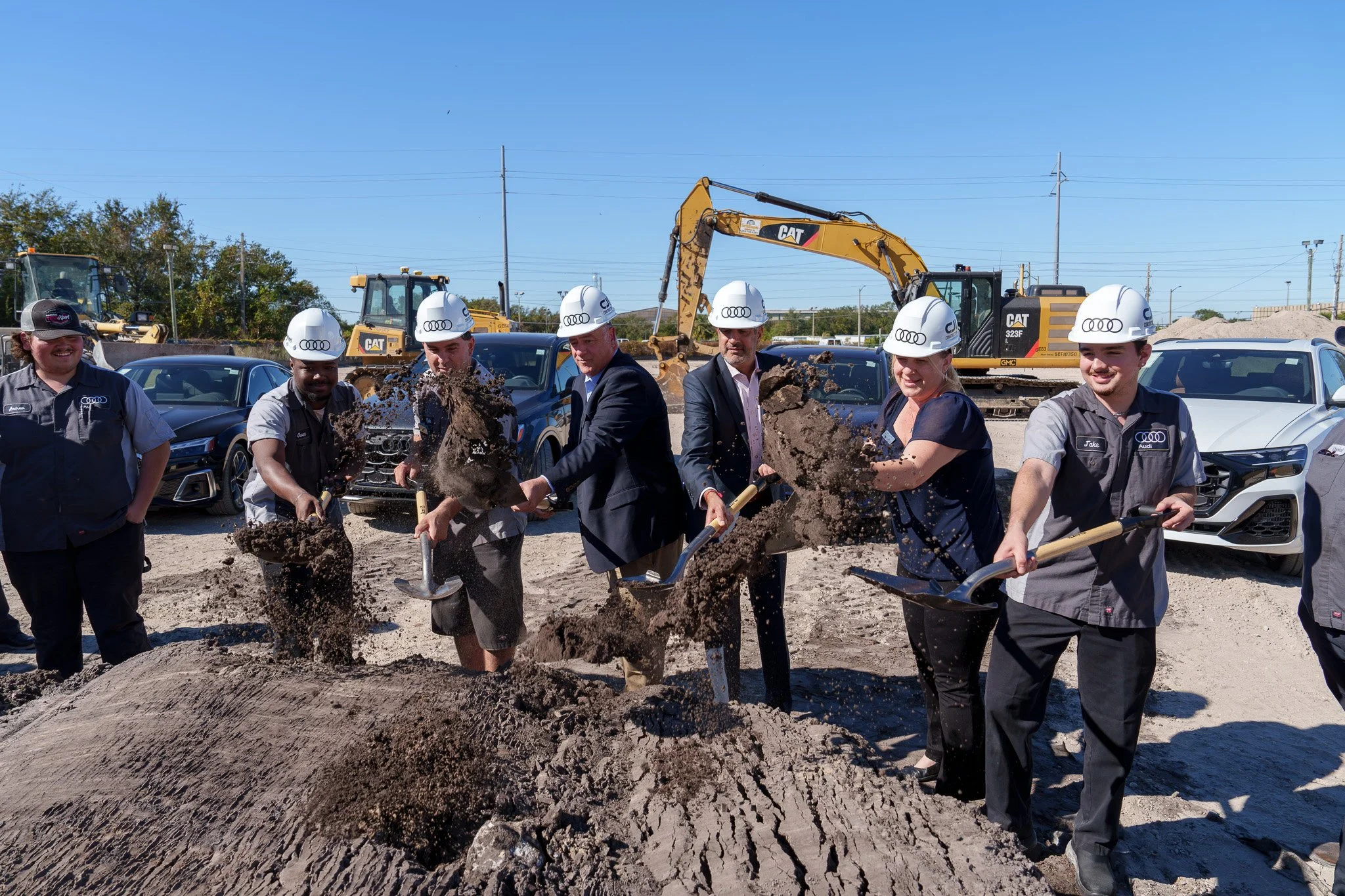 Group of people in hard hats and business attire standing around a large pile of dirt or mud at a construction site, with excavators and cars in the background.