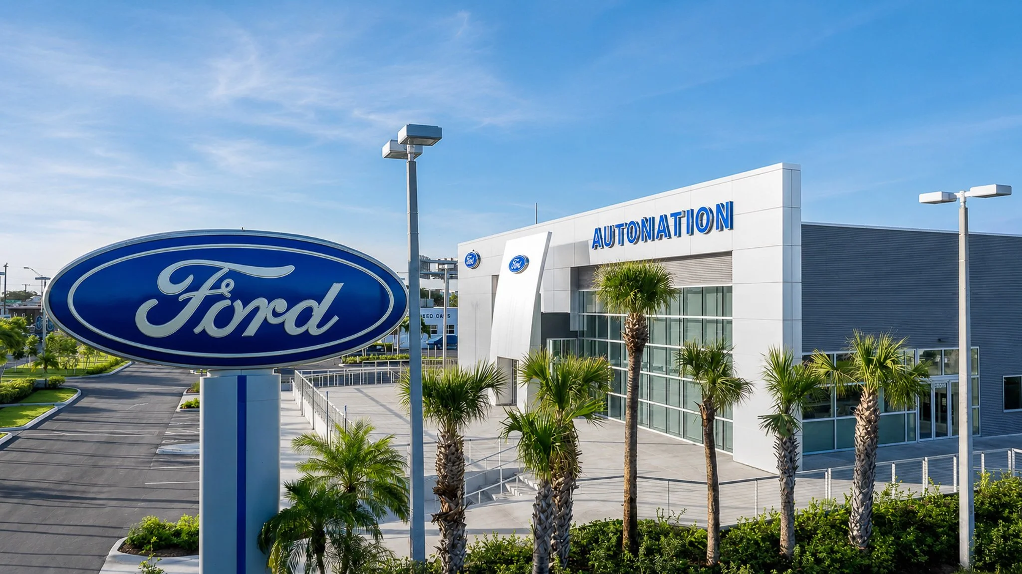 Exterior view of a Ford dealership with a large Ford logo sign in the foreground and a Modern building with blue 'Autonation' sign. Palm trees and cloudy blue sky are visible.