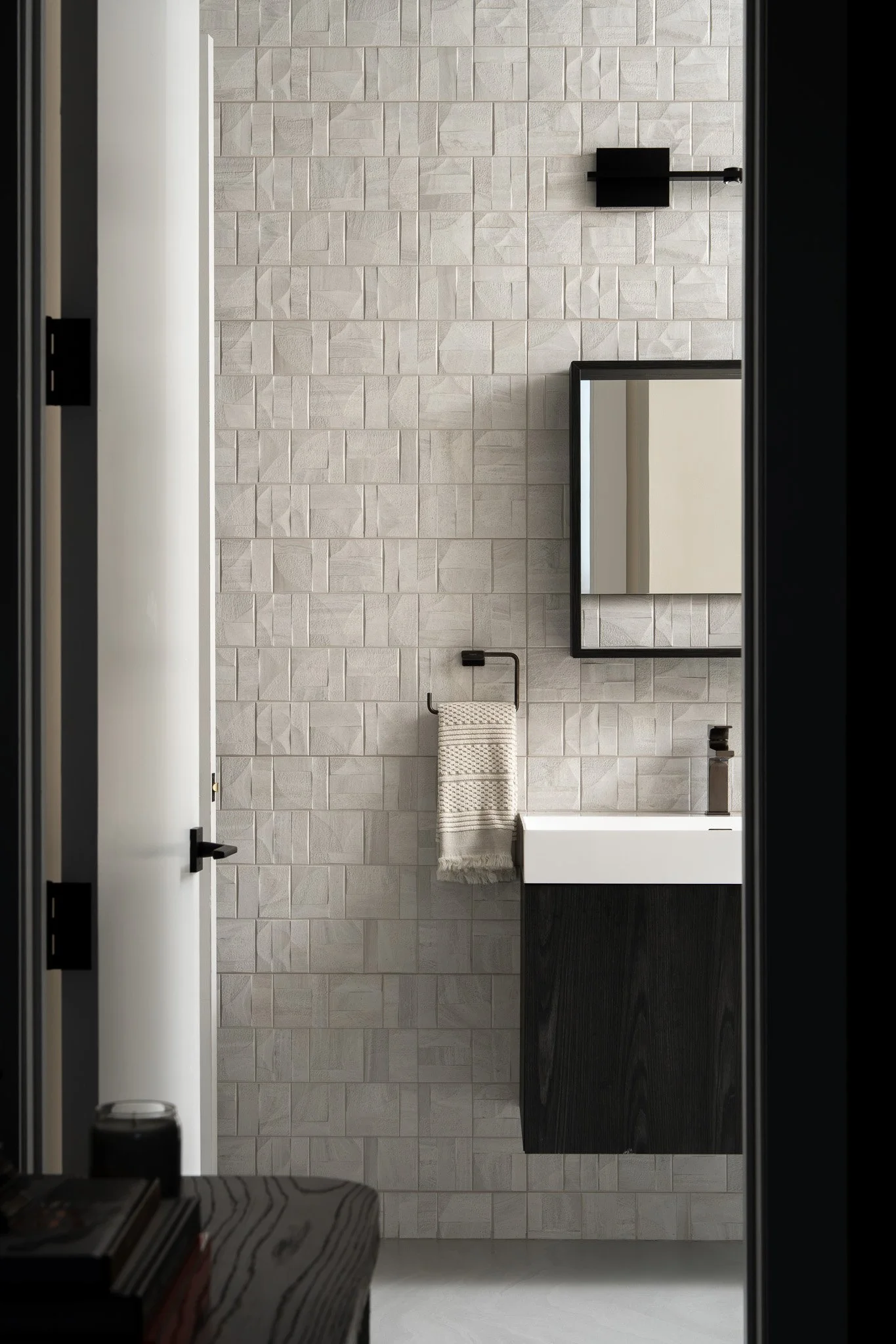 A modern bathroom with a textured beige tile wall, a black framed mirror, a black sink vanity, a towel hanging on a black rail, and a small black wall lamp. A black door frame is visible on sides, and a striped fabric is partially seen in the foregro