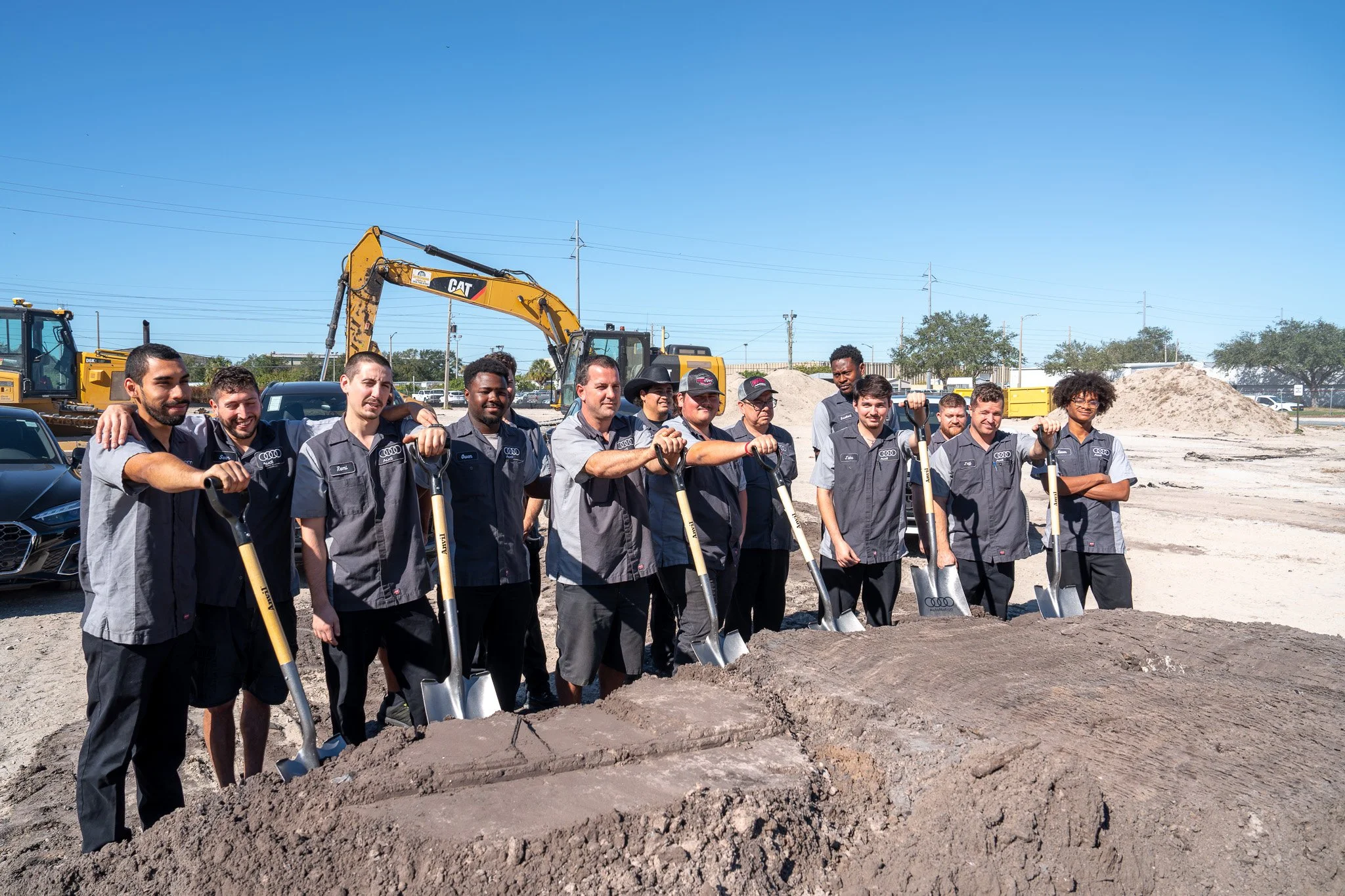 A group of construction workers, mostly men, standing with shovels at a construction site with dirt and machinery in the background under a clear blue sky.
