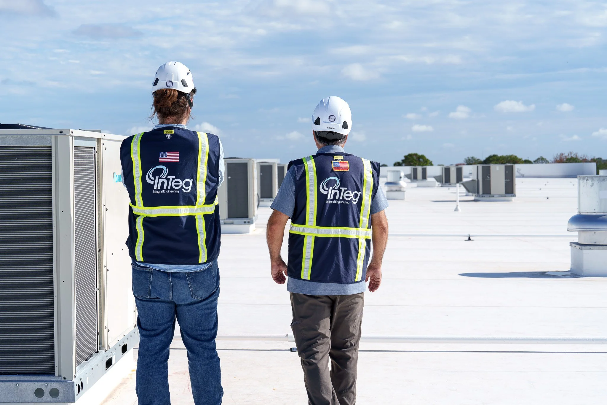 Two technicians wearing safety vests and helmets inspect HVAC units on a roof under a partly cloudy sky.