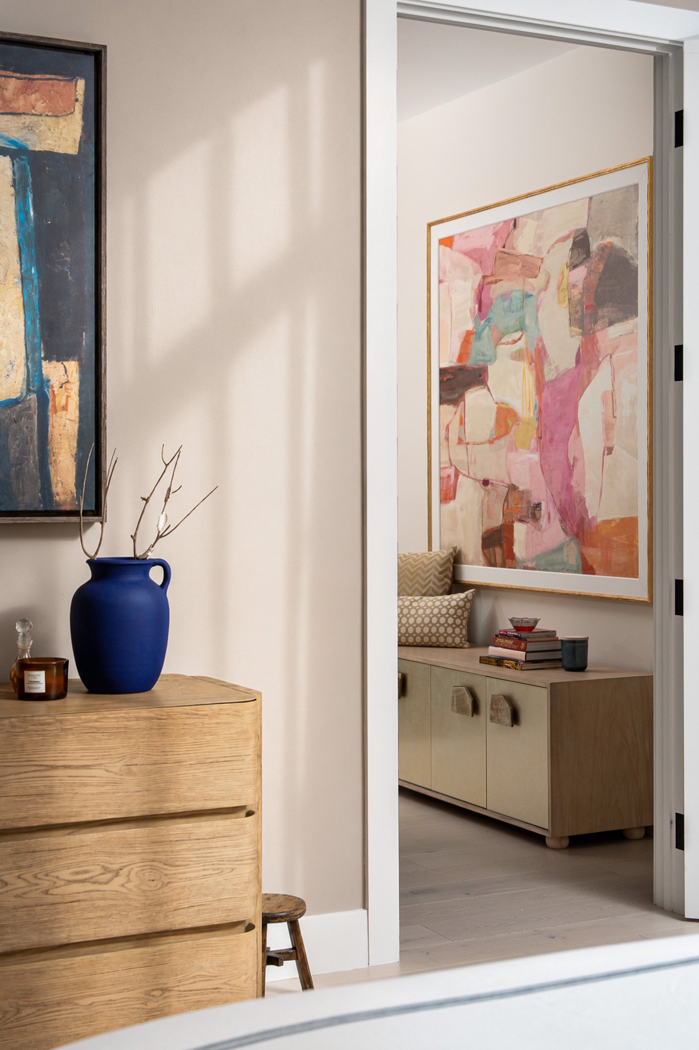 A view from a room showing a wooden dresser with a blue vase holding branches, and a small candle jar. In the adjacent room, a picture with abstract art hangs above a light-colored cabinet decorated with books, a bowl, and a black cup.